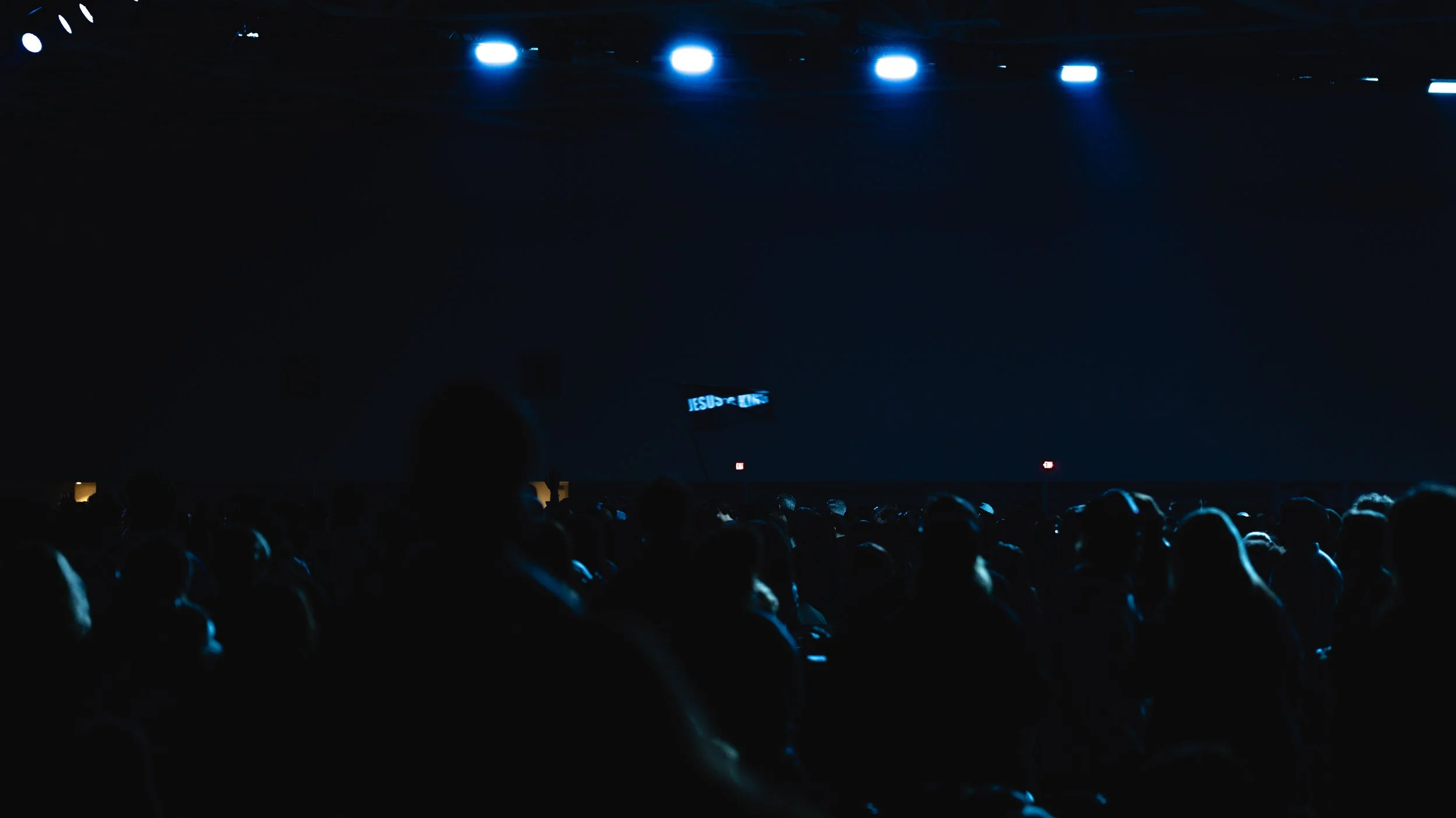 Silhouette of a crowd of people seated in a dark auditorium with blue stage lights overhead and a small illuminated sign that reads 'JESUS' in the background.