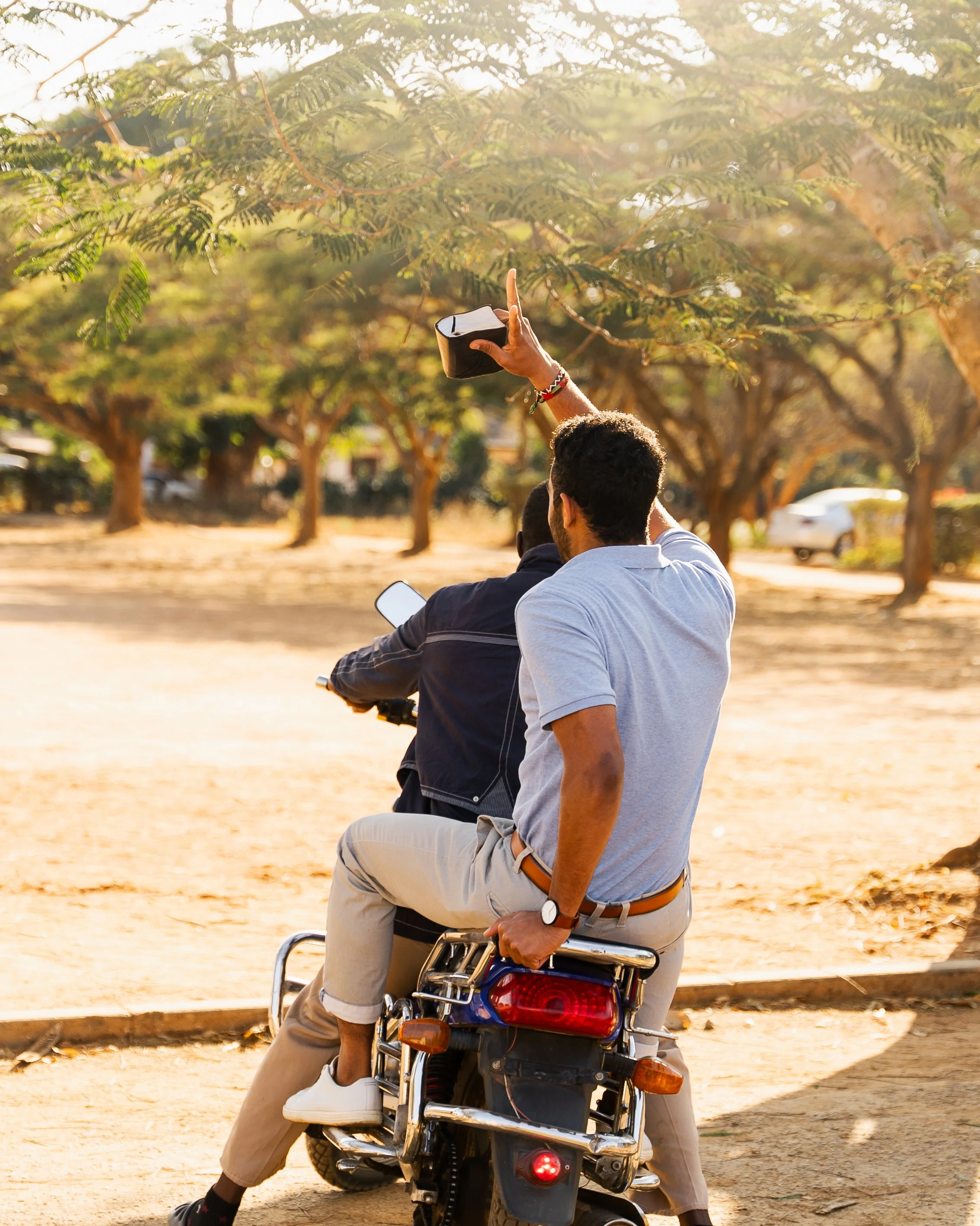 Two young men are riding a motorcycle on a dirt path, with one of them holding a smartphone and the other using a smartphone, surrounded by trees in a park or outdoor setting.