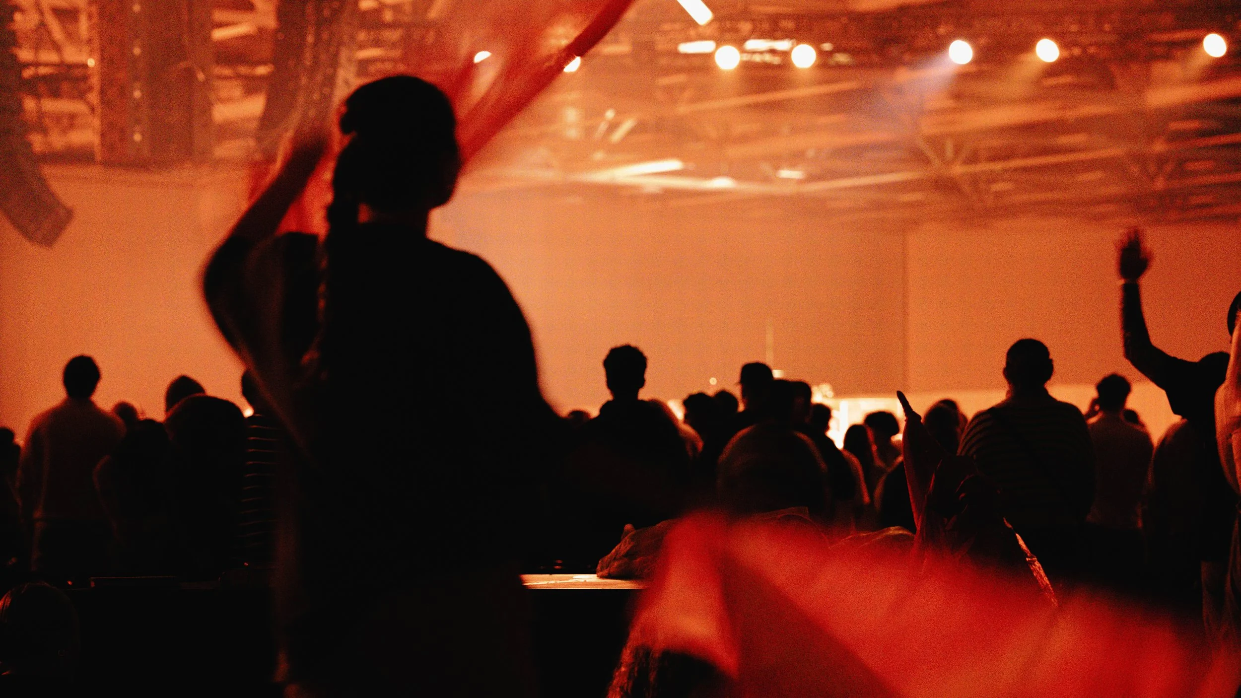 People gathered in a dimly lit indoor venue with warm orange lighting, some with hands raised, silhouetted against the background. A woman with braided hair is in the foreground, facing the stage or performance area.