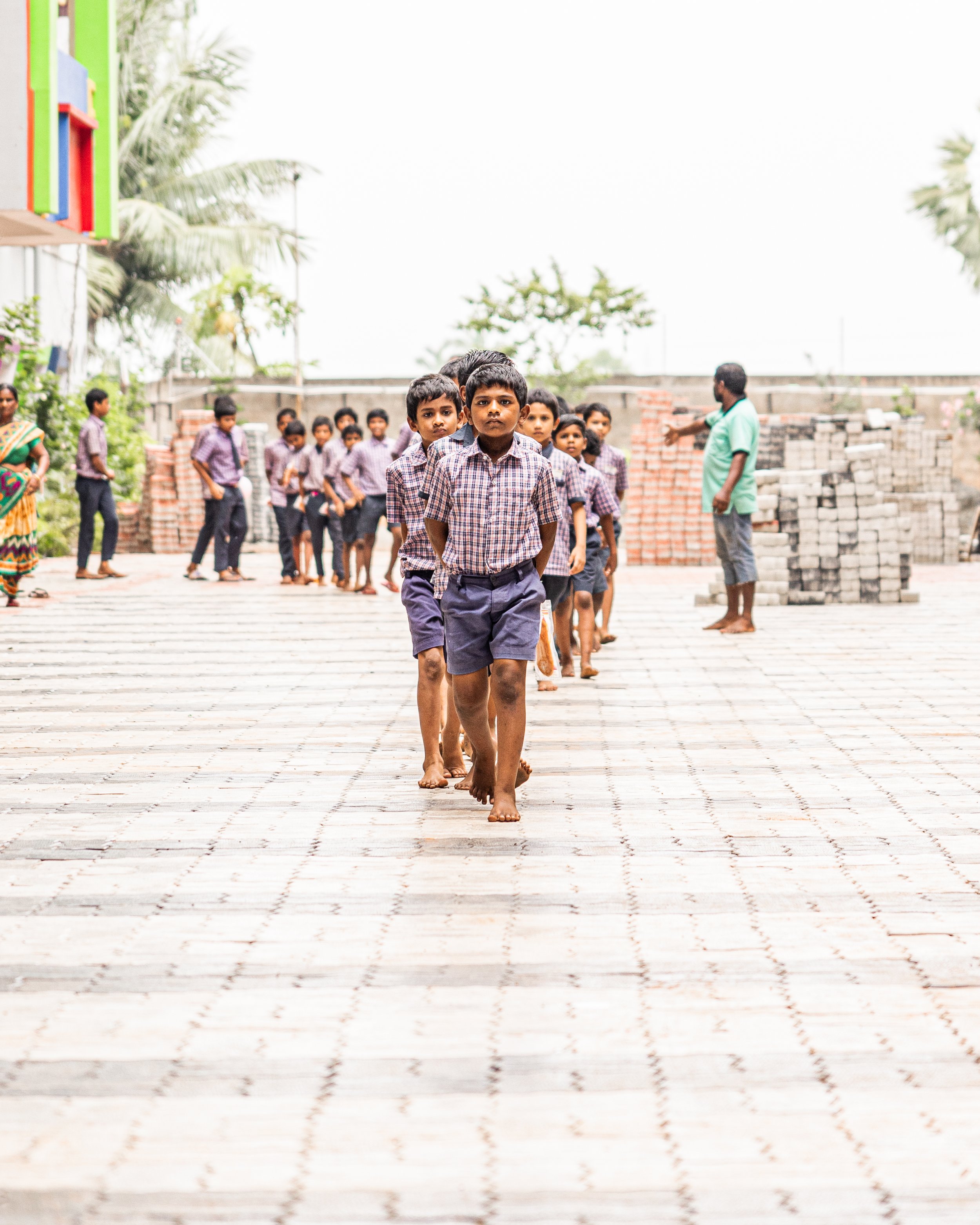 Children in school uniforms standing in a line outdoors, with teachers or adults nearby, on a paved area with construction materials in the background.