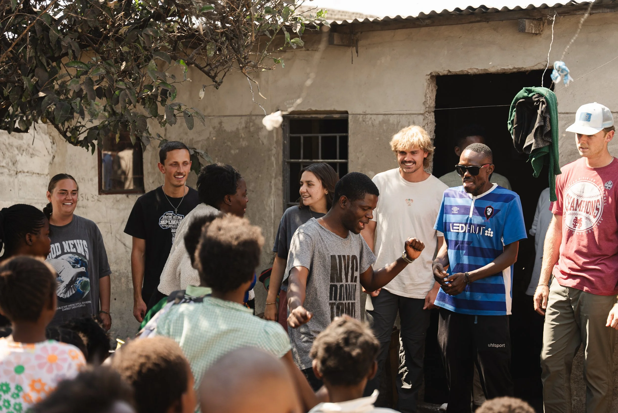 A group of people, including a mix of local residents and visitors, gathered outside a rustic building, engaging in a lively outdoor activity or celebration, with some smiling and interacting.
