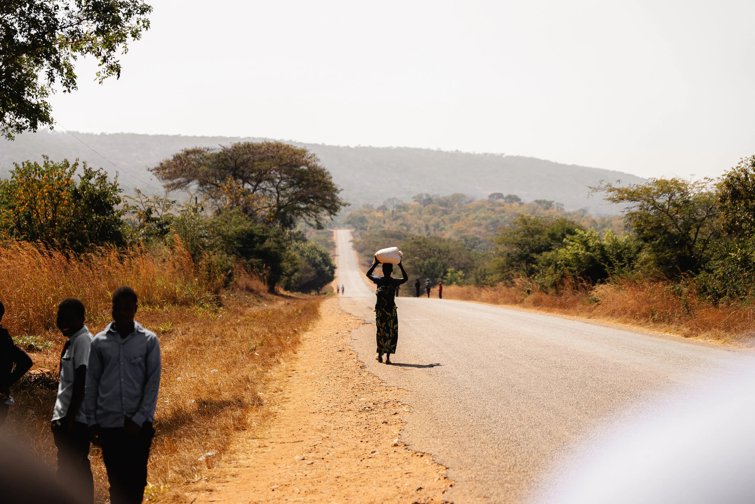A woman walking along a rural road carrying a white object on her head, other people walking further down the road, and children standing on the side of the road, with dry grass and trees on either side and hills in the background.
