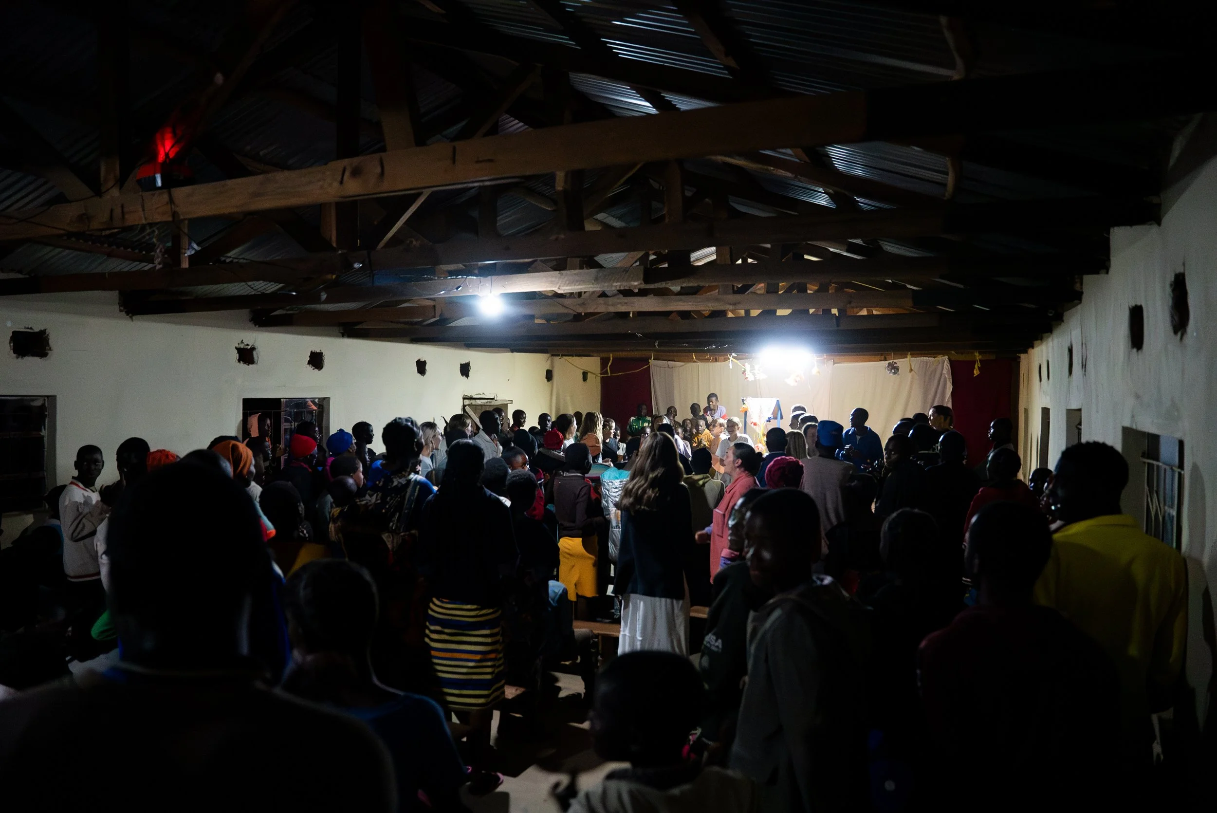 A large group of people gathered inside a building with wooden beams and a corrugated metal roof. The crowd appears to be attending a community event or meeting, with some individuals standing and others seated. There is a bright light illuminating t