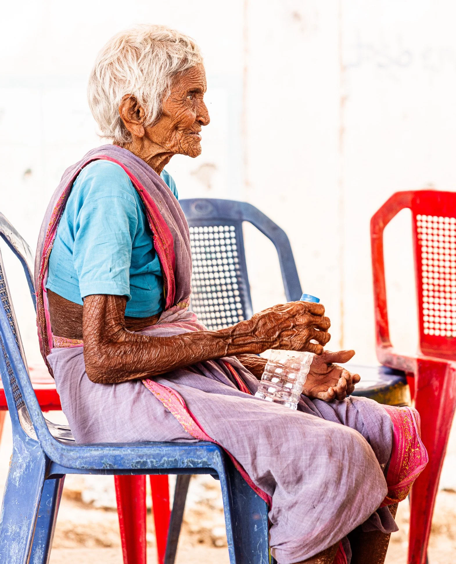 An elderly woman with gray hair and dark, wrinkled skin, wearing a teal blouse and a patterned pink sari, sitting on a blue plastic chair holding a clear water bottle with a blue cap, surrounded by red plastic chairs.