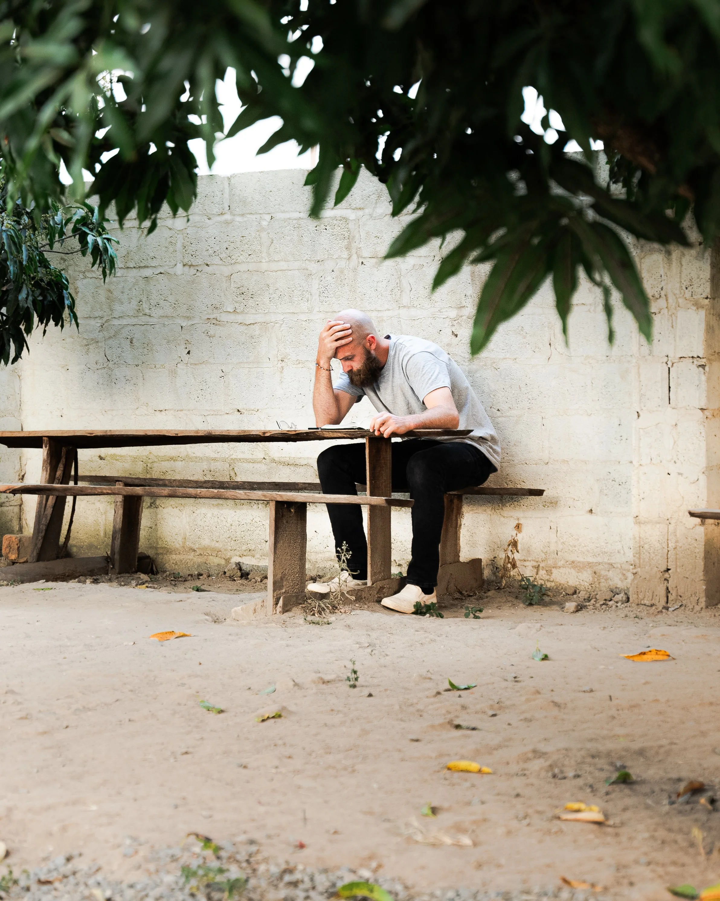 A man sitting on a wooden bench outside, looking distressed with his hand on his forehead, beneath overhanging green leaves and a beige brick wall.