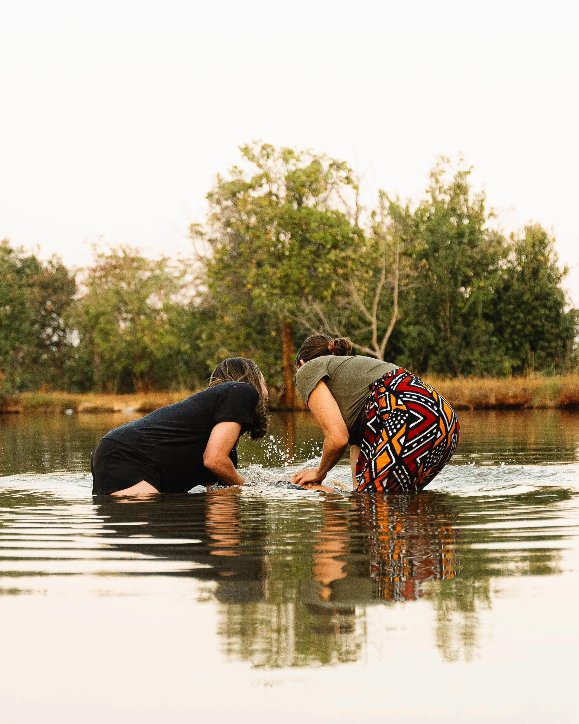 Two women standing in a river, leaning forward and touching the water, with trees in the background during sunset.