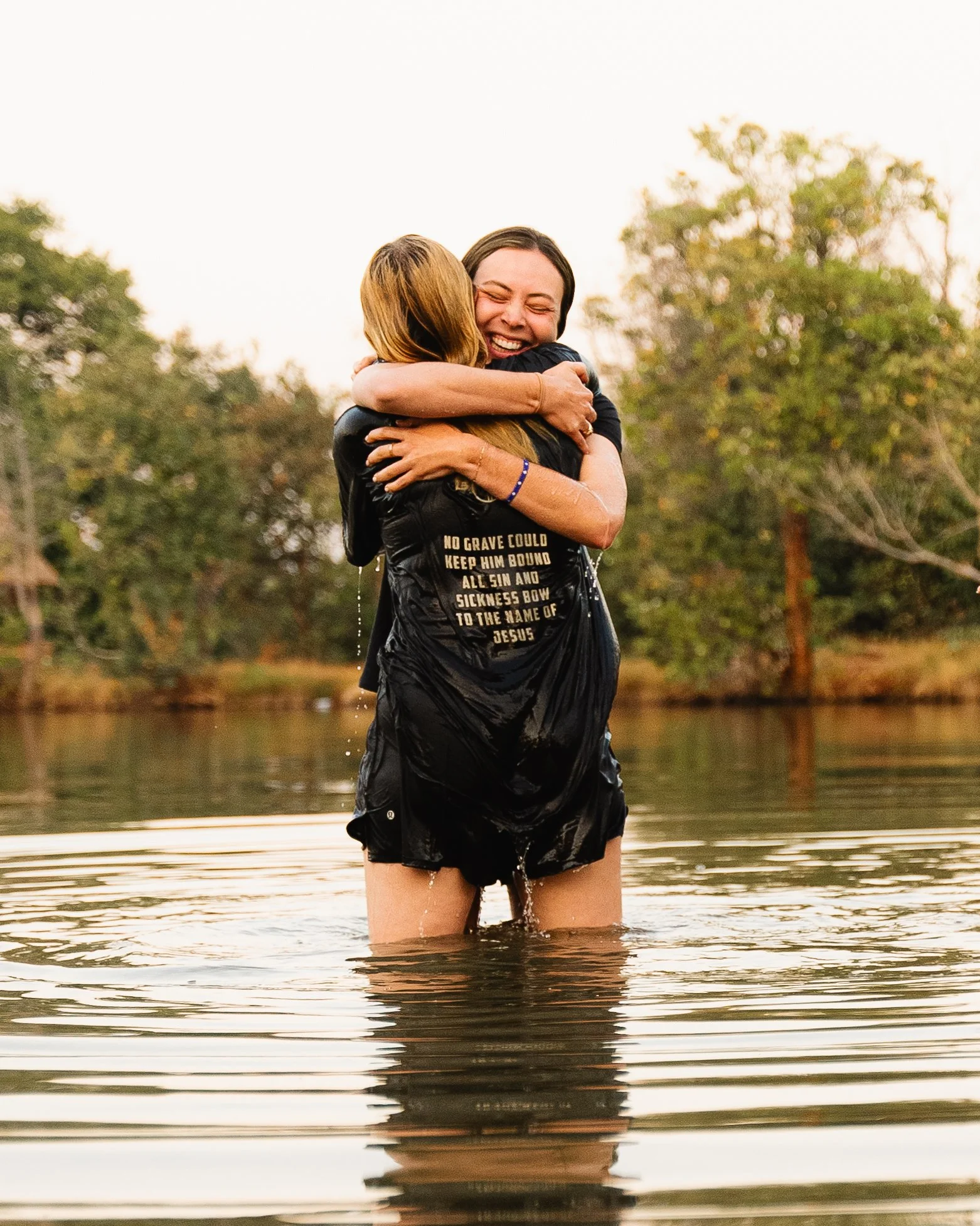 Two women joyfully hugging in a lake, with trees and a cloudy sky in the background.