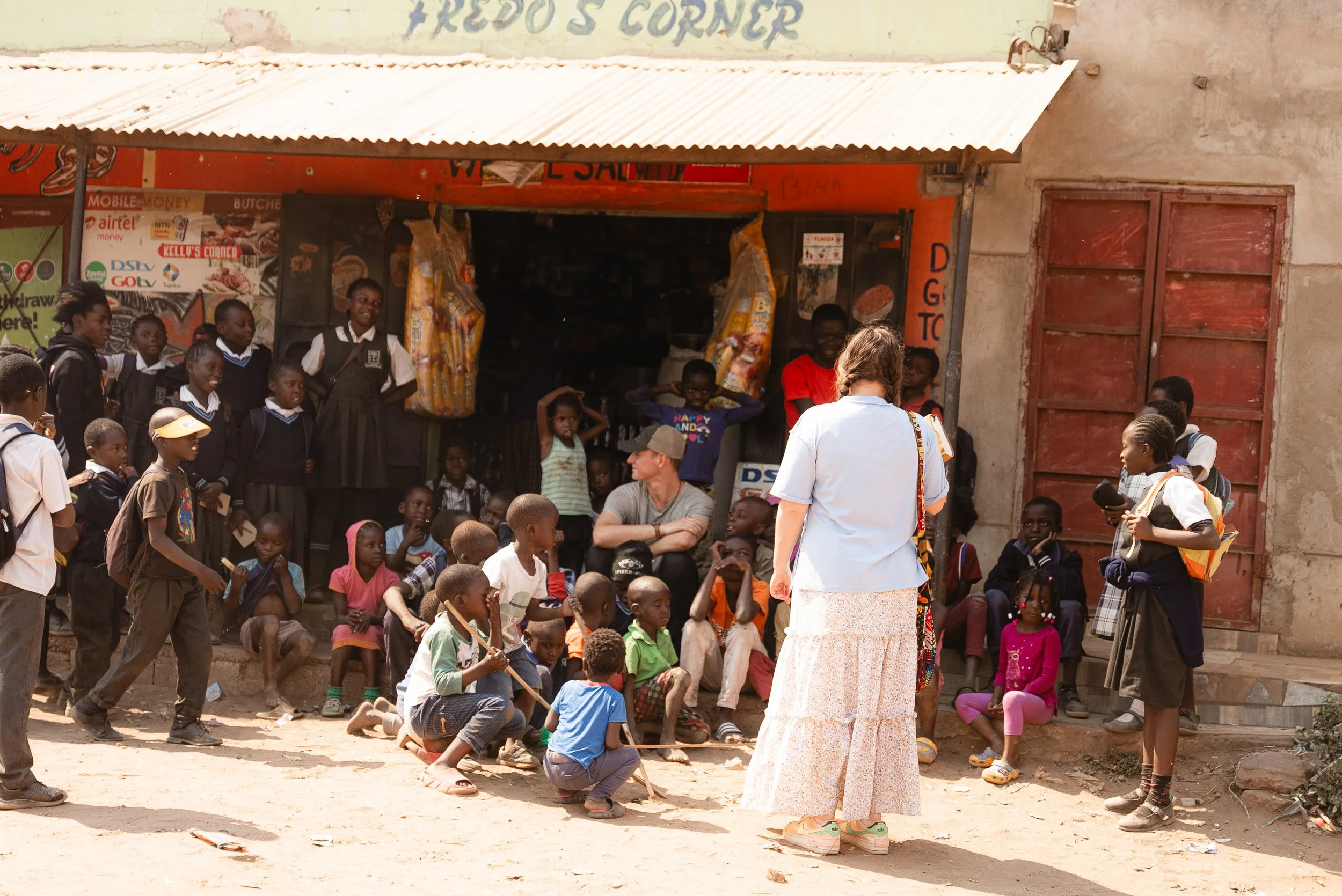 A group of children and adults gathered outside a small shop on a dusty street, with some sitting, standing, or playing. The shop has an orange facade with signs and advertisements, and people are engaged in conversation and activities outdoors.