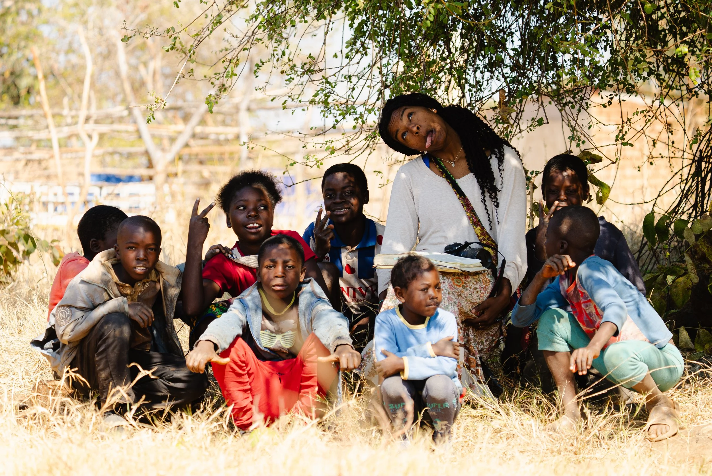 A group of children and a woman, some sitting and some squatting, outdoors under a tree on grassy ground, making playful gestures and facial expressions.