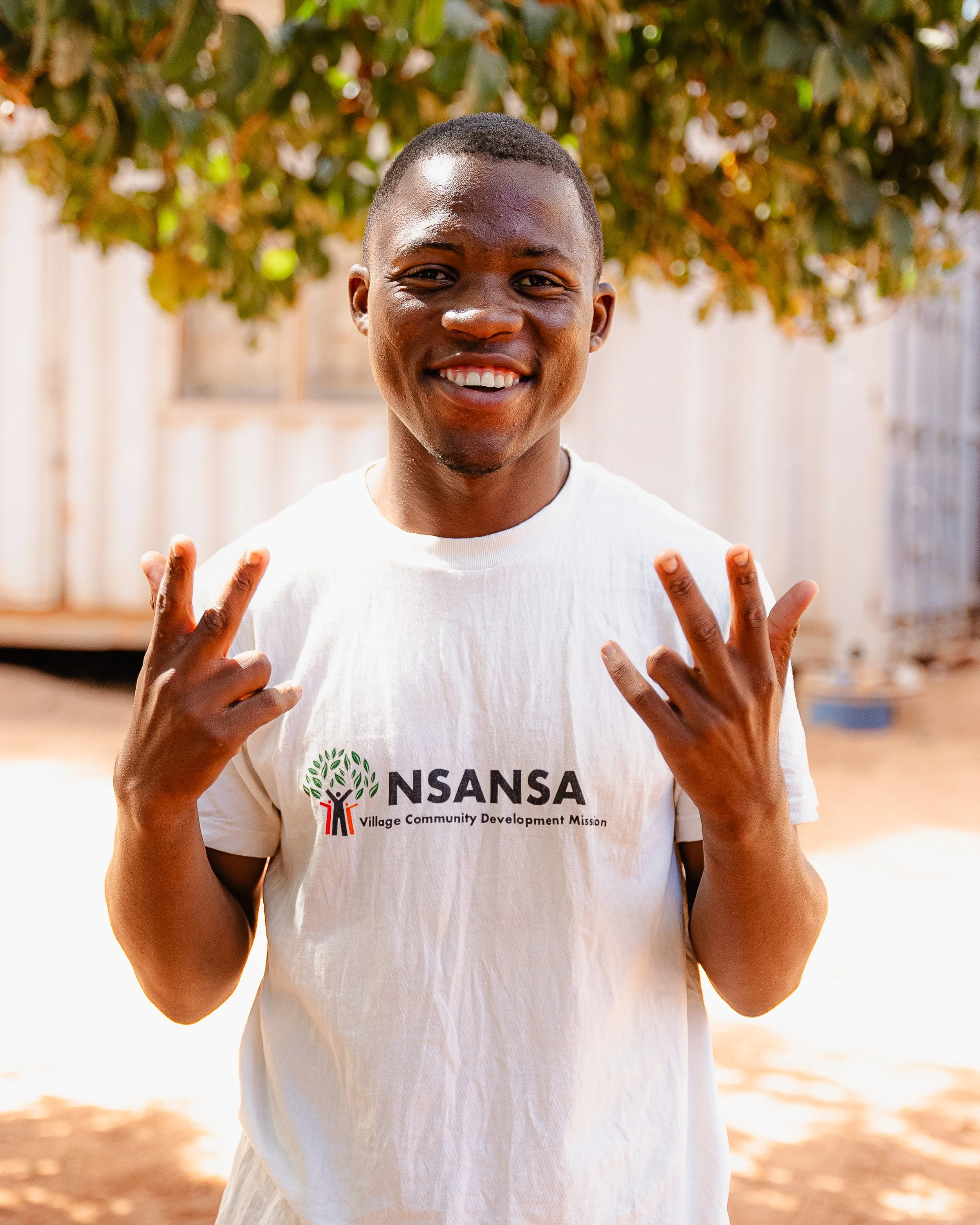 A young man smiling outdoors, making peace signs with both hands, wearing a white T-shirt with the logo and text for NSANSAN Village Community Development Mission, standing in front of a tree and blurred background.