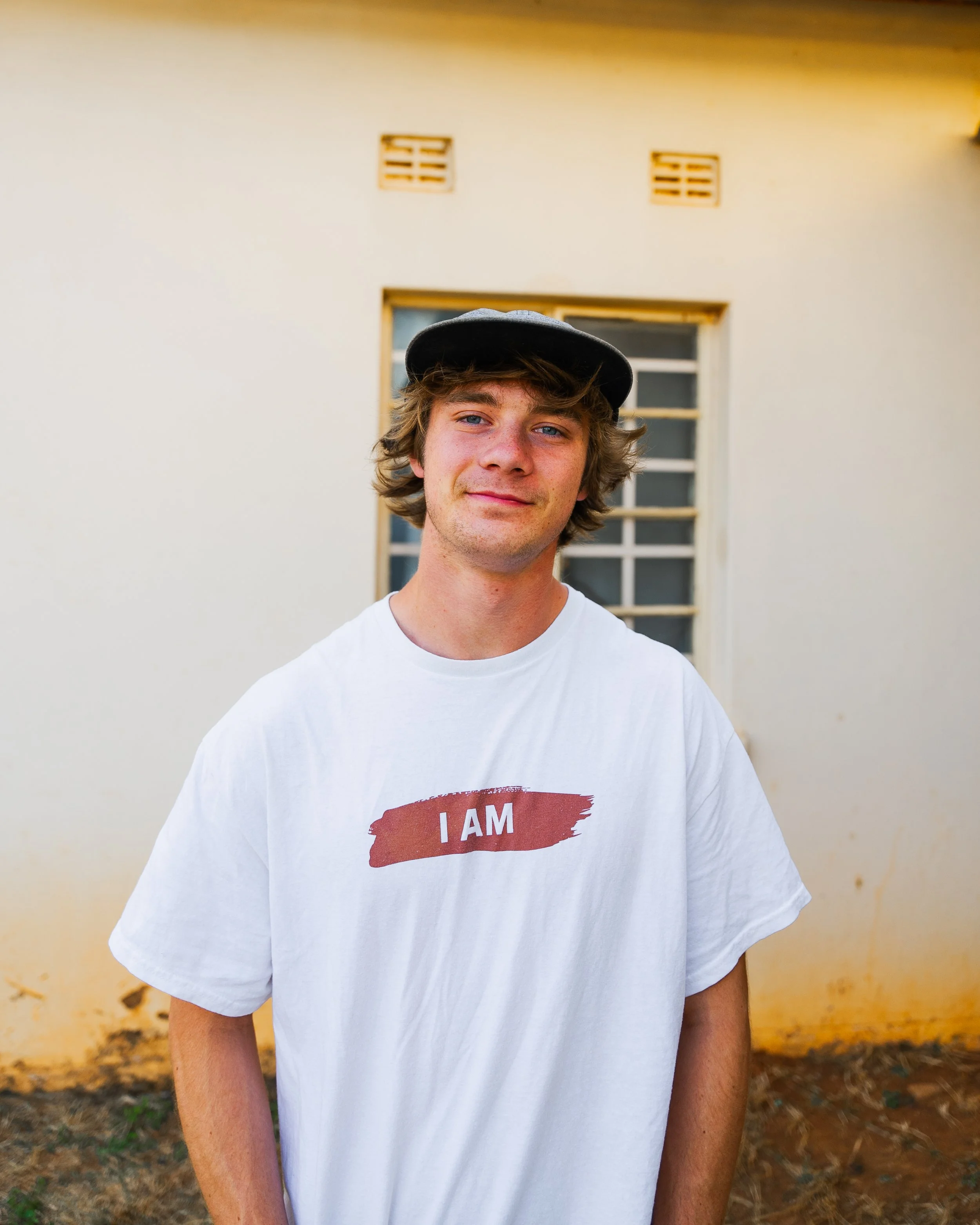 A young man with blond hair, wearing a black cap and a white T-shirt that says 'I AM' in red. He is standing outdoors in front of a white wall with a window.