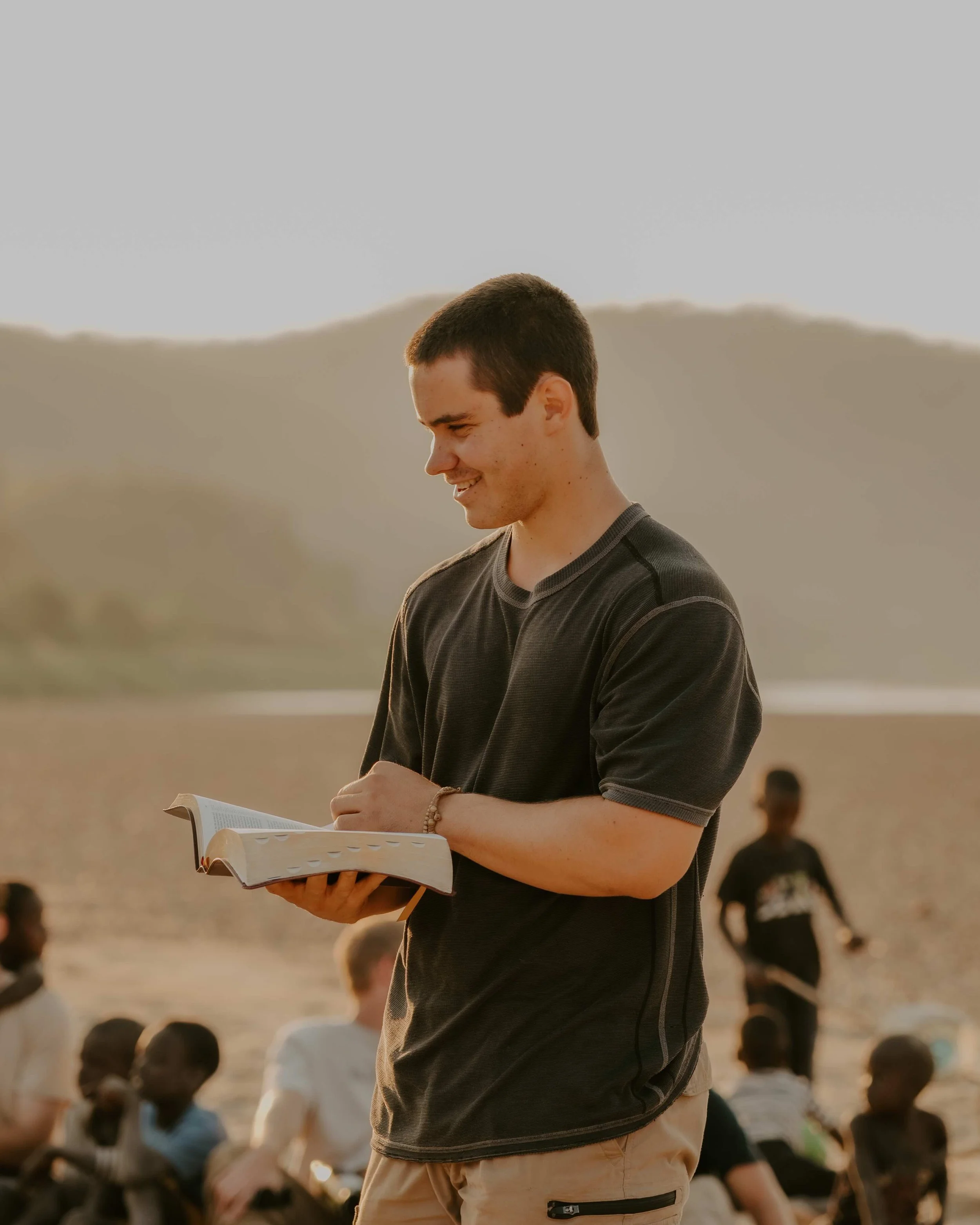 A young man with short dark hair smiling while reading a book outdoors during sunset, with group of children sitting on the ground in the background.