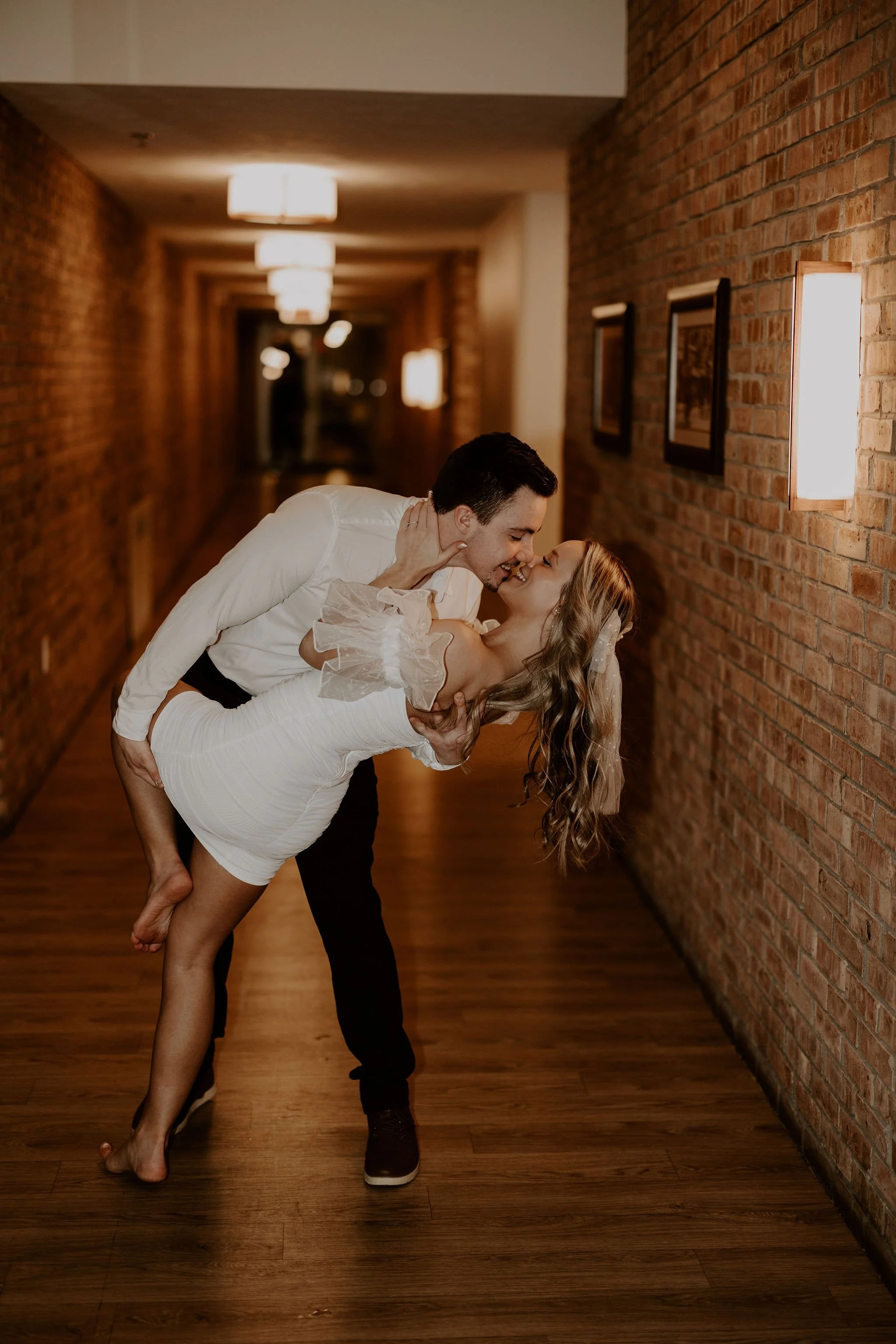 A man and woman are dancing together in a hallway with brick walls and wooden floors. The man is leaning over and holding the woman as she leans back, smiling and looking at him. The lighting is warm and soft.