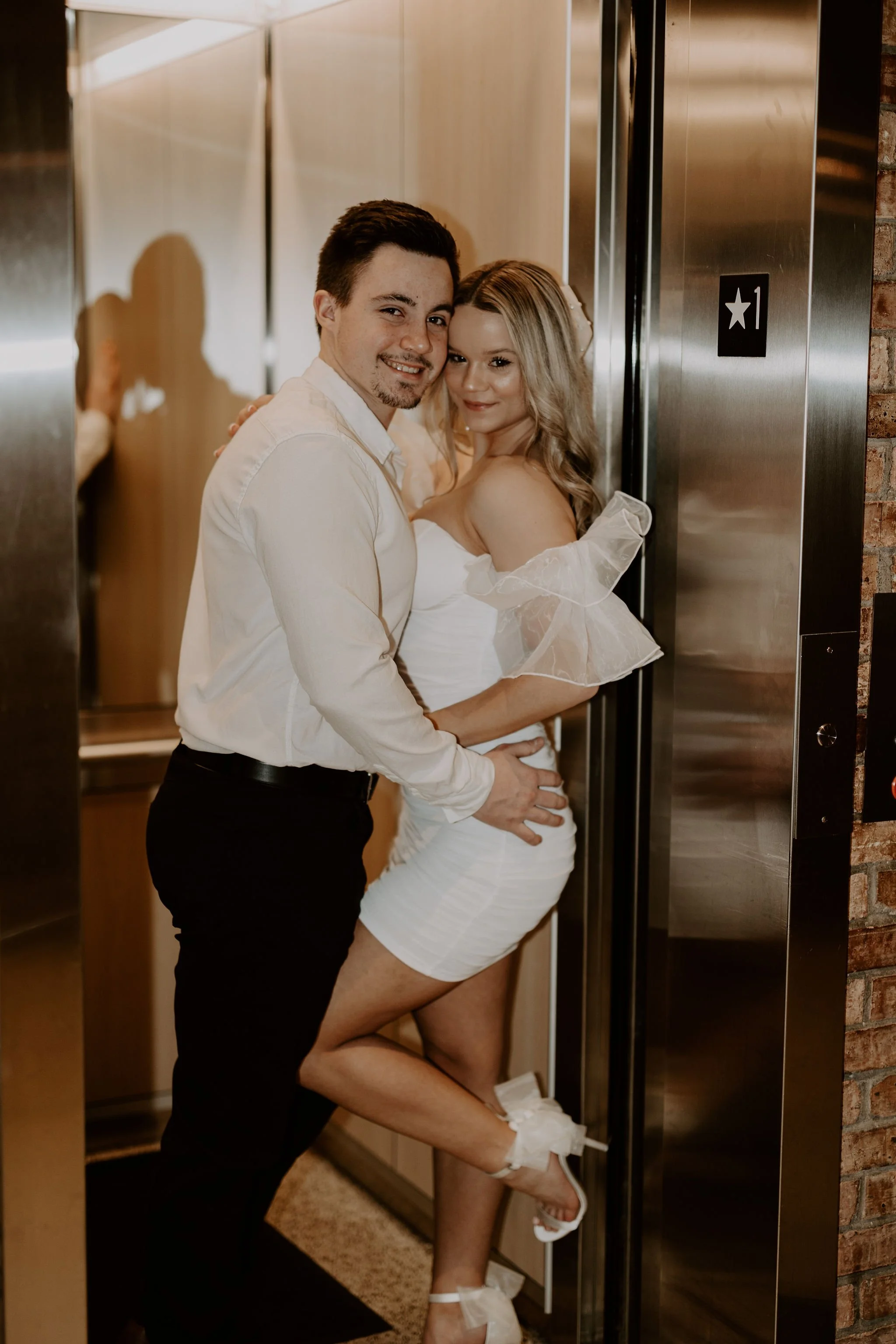 A smiling young couple embraces in front of an elevator door, with the woman wearing a white dress and the man in a white shirt and black pants.