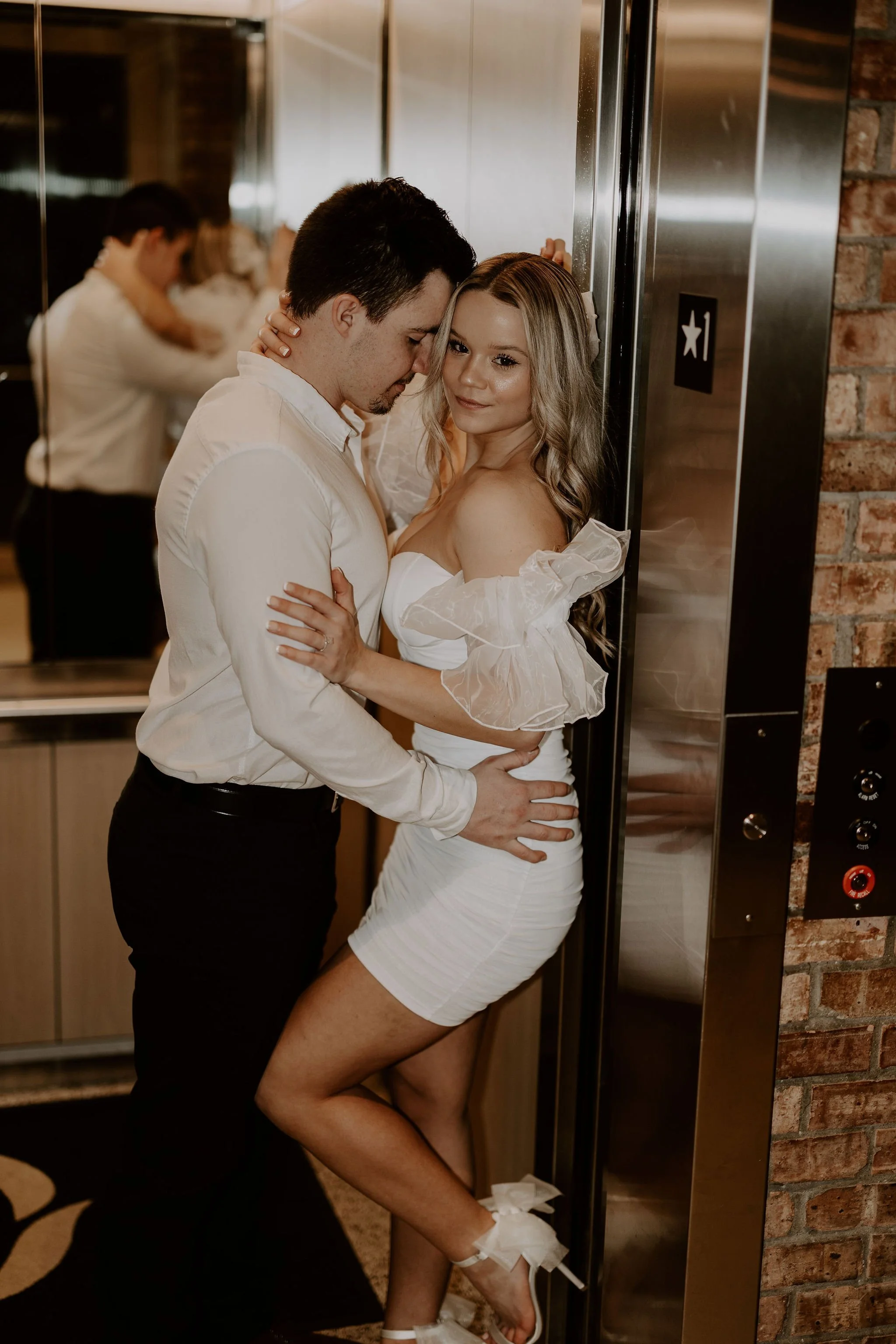 A young man with dark hair and a woman with long, wavy blonde hair are standing close together in an elevator. The man is leaning his forehead against the woman's temple with his eyes closed, and she is smiling softly at the camera. They are dressed 