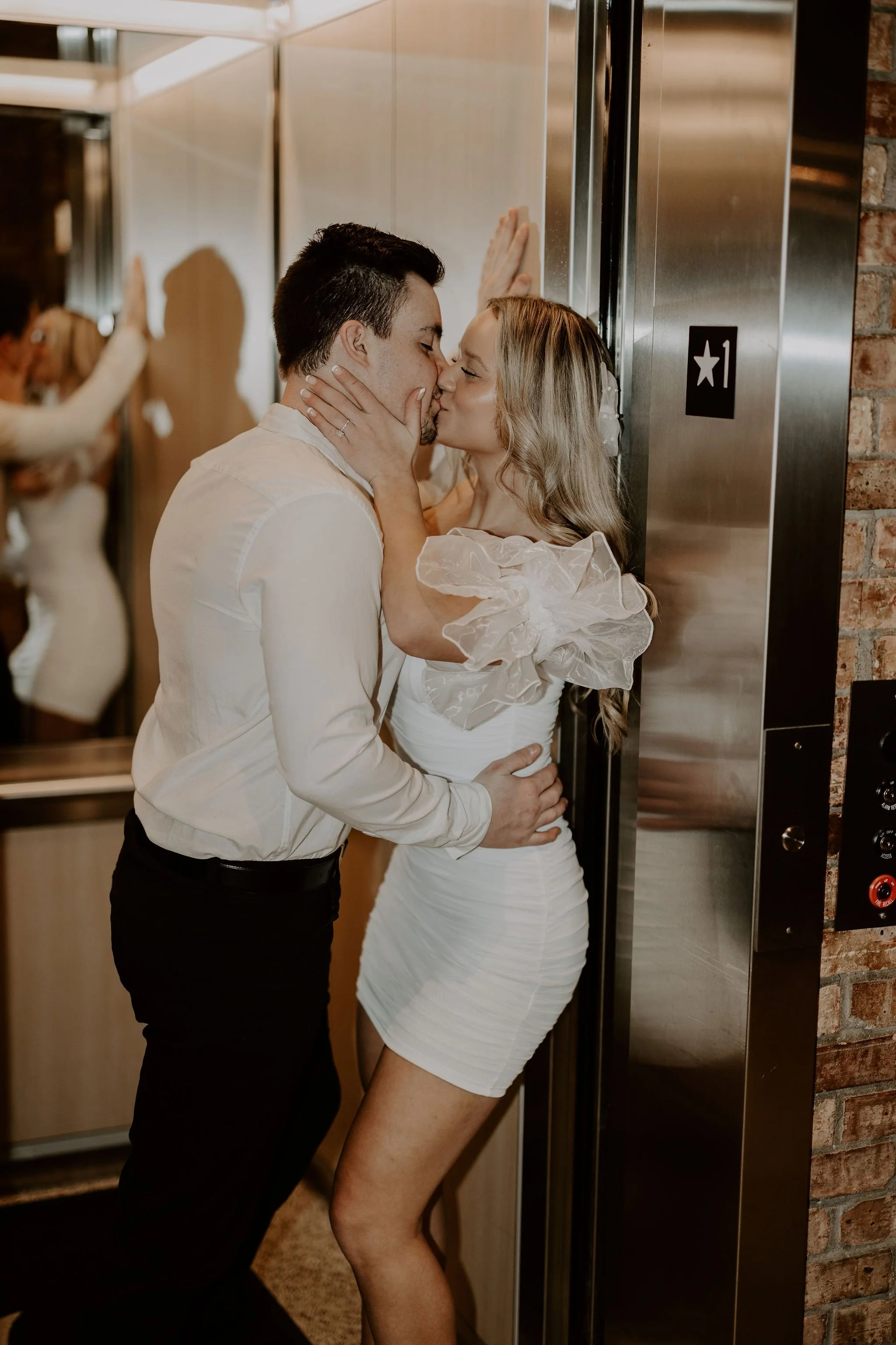 A man and woman kissing in an elevator, with the woman wearing a white dress and the man in a white shirt. The woman has her hands on the man's face, and the man has one hand on her waist. The elevator has a stainless steel wall with a black panel sh