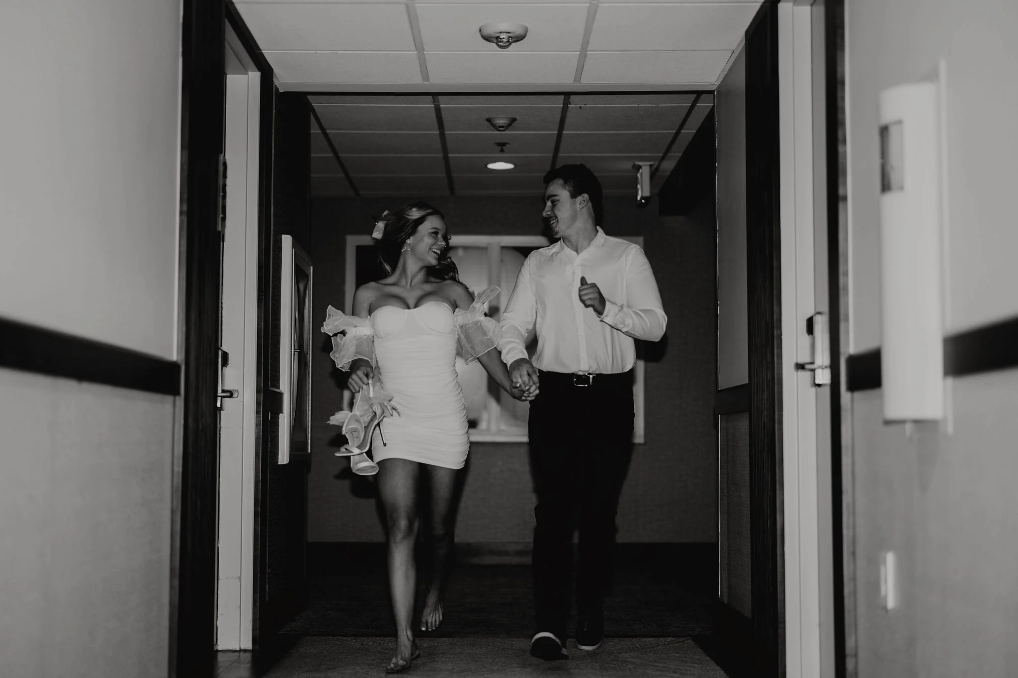 A black-and-white photo of a couple walking through a hallway, holding hands, smiling, and looking at each other. The woman is in a off-shoulder dress and holding a pair of high-heeled shoes.