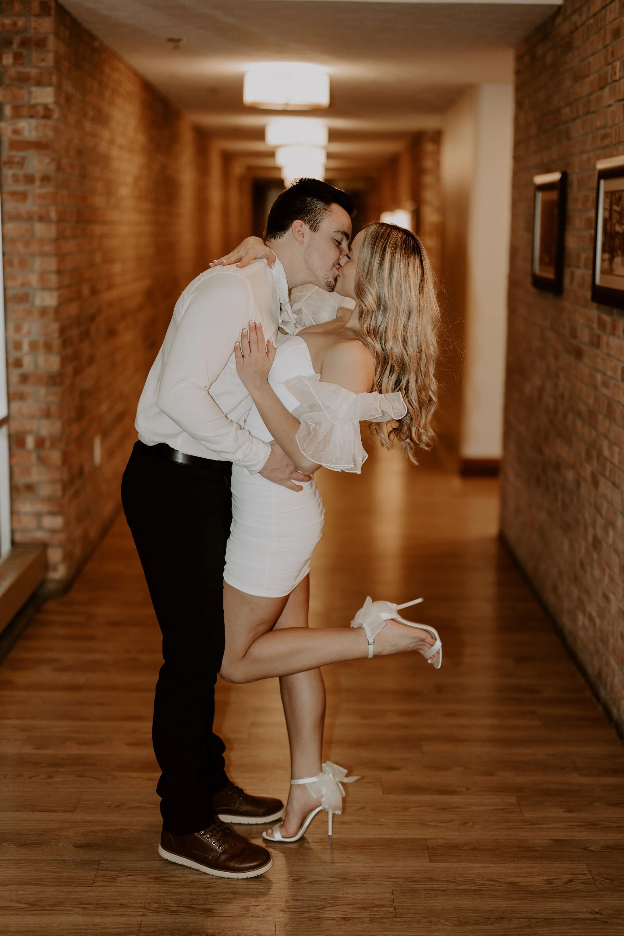 A couple kissing in a hallway with brick walls. The woman, dressed in a white off-shoulder dress, is kneeling on one knee and wearing white high-heeled shoes with bows. The man is standing, wearing a white shirt and black pants, with his arms around 
