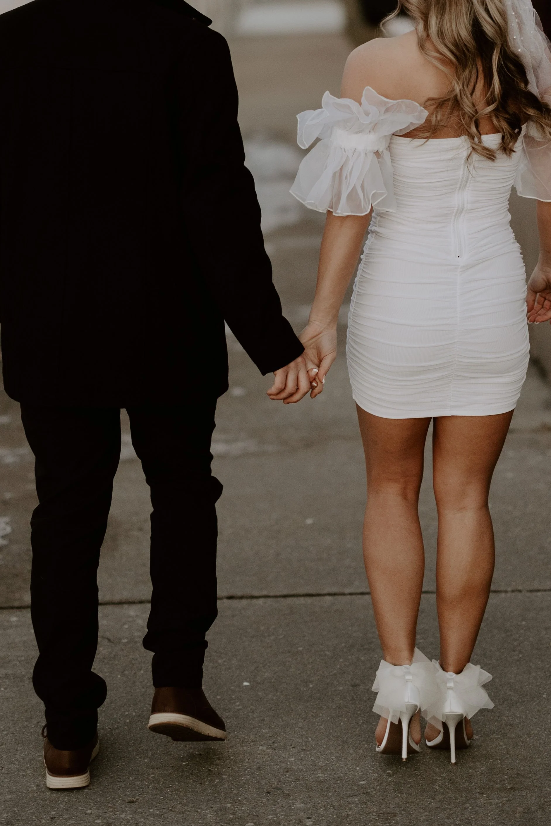 Close-up of a couple holding hands walking on a street, with the woman wearing a white, ruffled dress and high heels, and the man in a black suit and casual shoes.