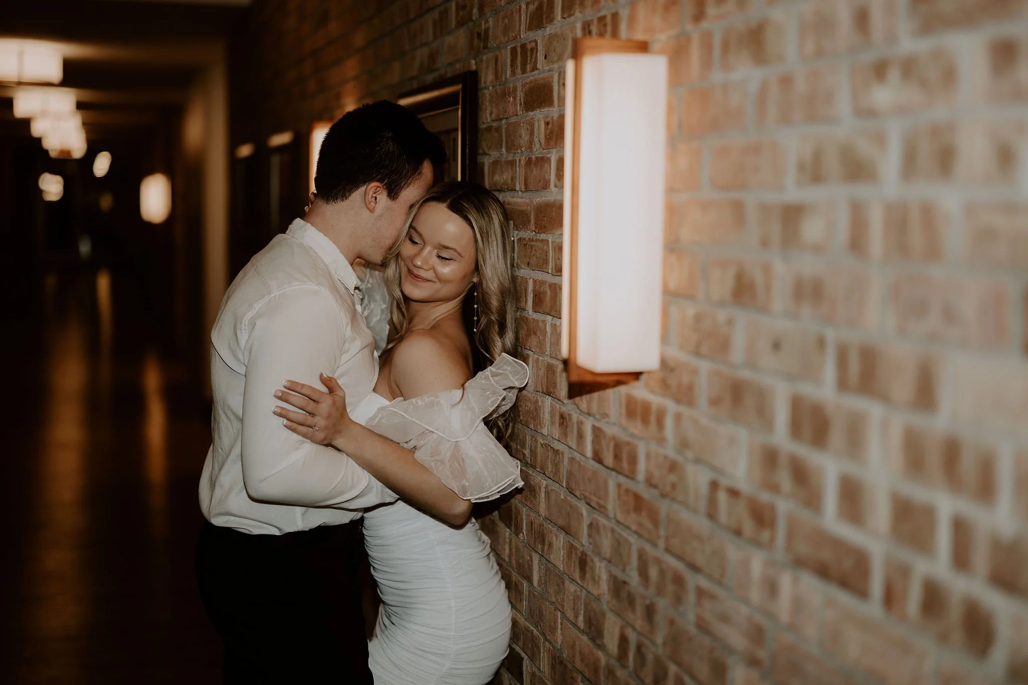 A couple embraces against a brick wall in a hallway with warm lighting, smiling and enjoying a romantic moment.