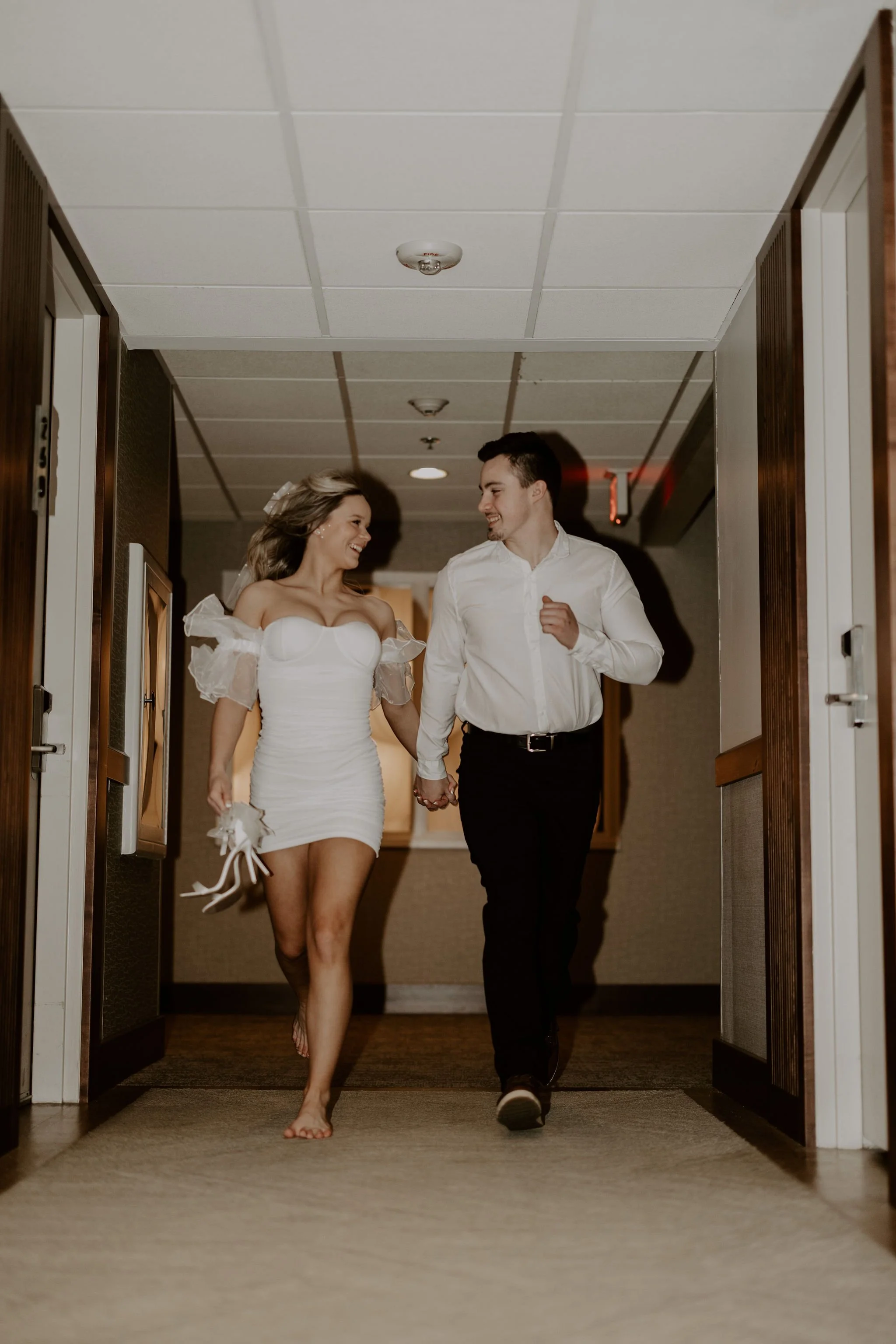 A young woman and man are walking hand in hand down a hallway, smiling and looking at each other. The woman is in a white dress with puffed sleeves, and the man is in a white shirt and black pants.
