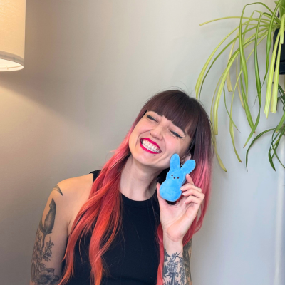 brigitte, a white woman with brown and pink hair and arm tattoos, squinting & smiling, holding a small blue bunny peep stuffed animal