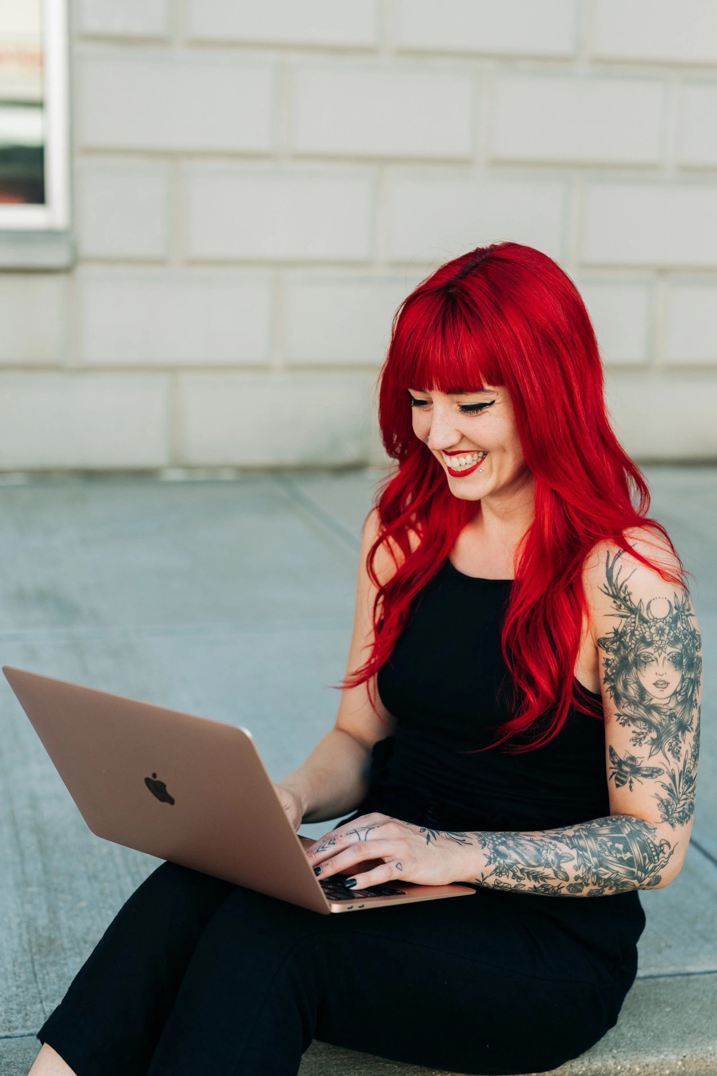 brigitte, a white woman with long curled bright red hair and arm tattoos, wearing black. sits outside on a business from steps smiling at her computer as she provides custom notion systems for small business owners