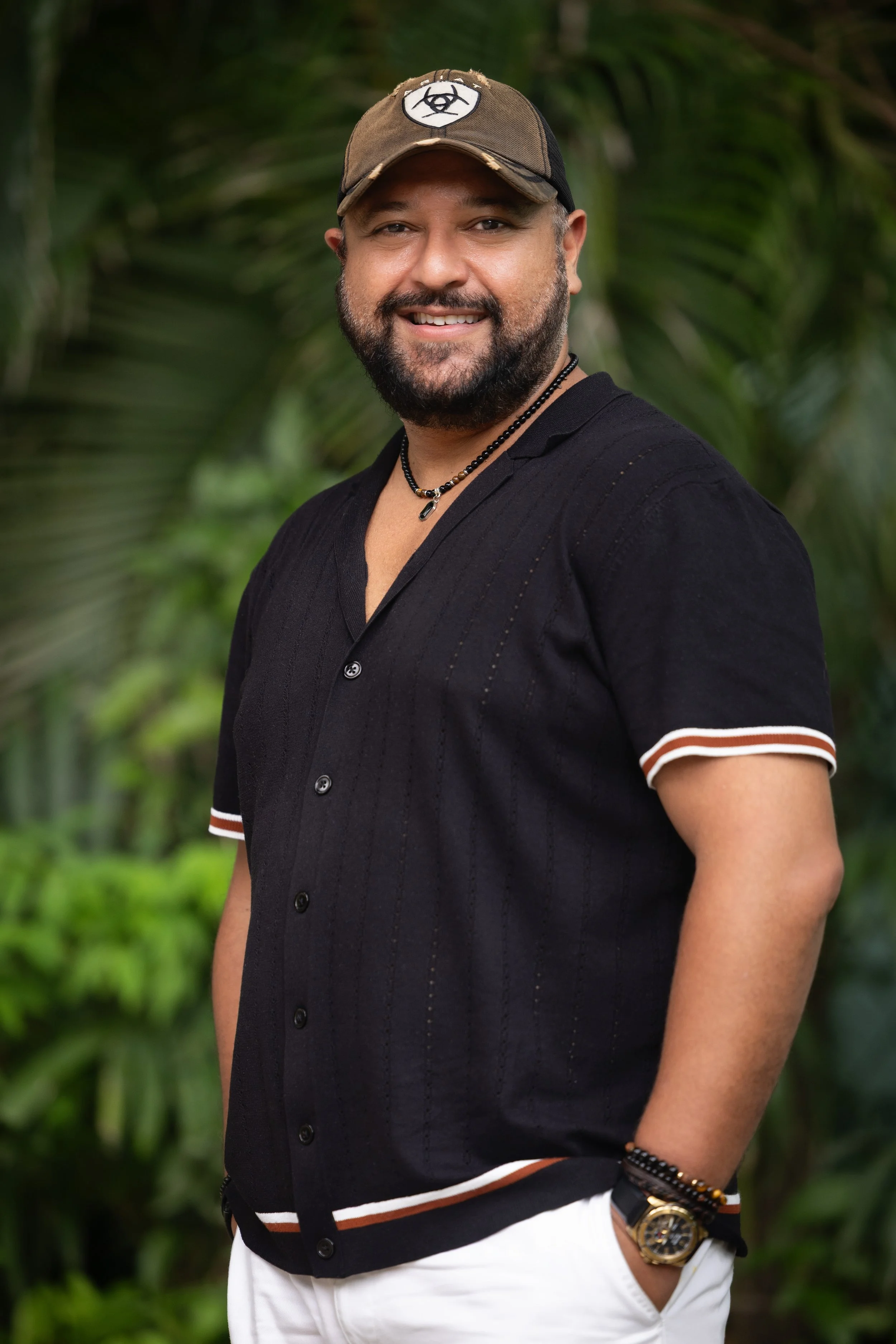 Un hombre sonriente con gafas, barba y bigote al aire libre entre exuberantes plantas tropicales.