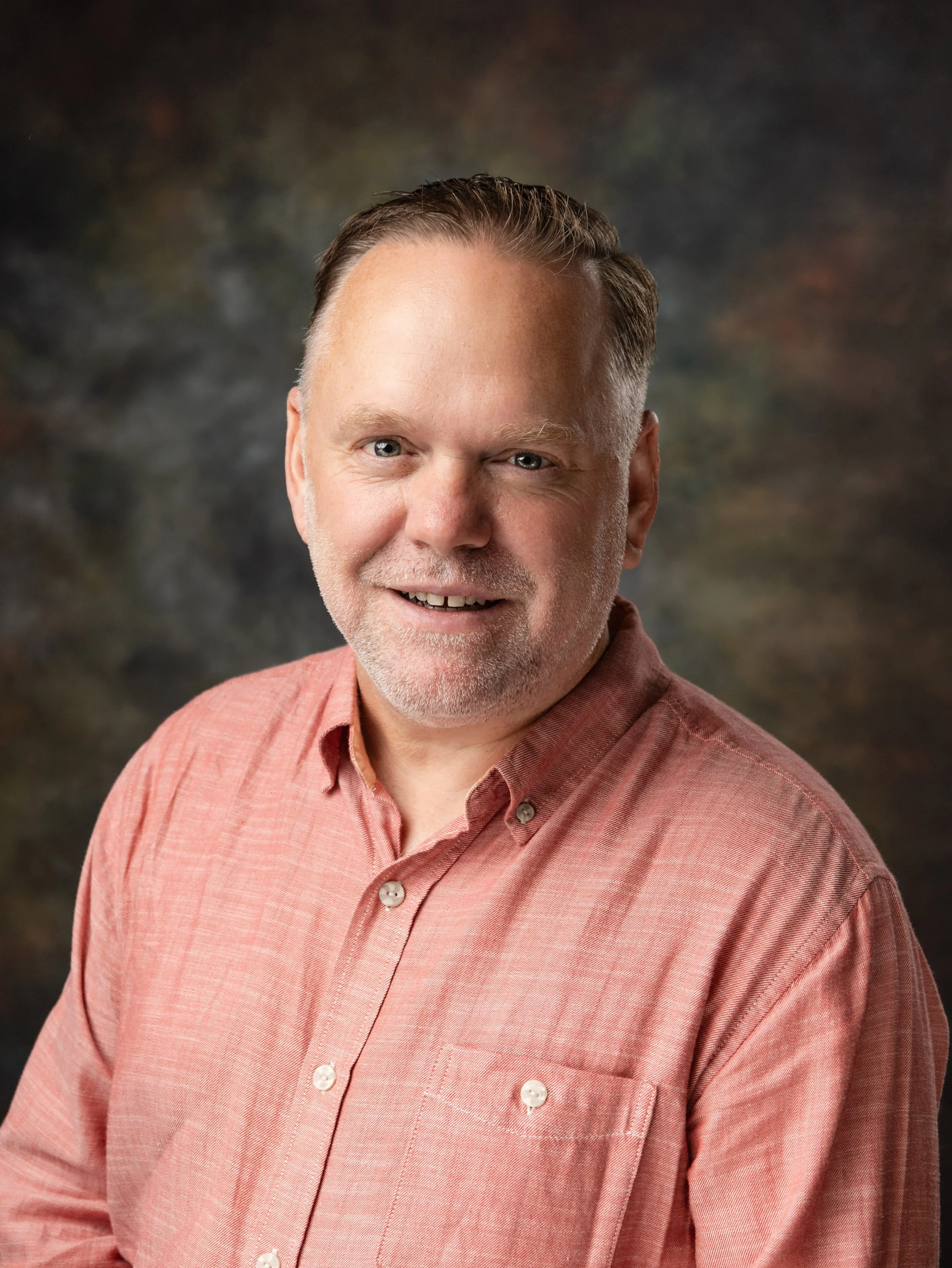 Close-up portrait of a smiling middle-aged man with short, dark hair, a beard, and wearing a light-colored button-up shirt, against a blurred brown background.