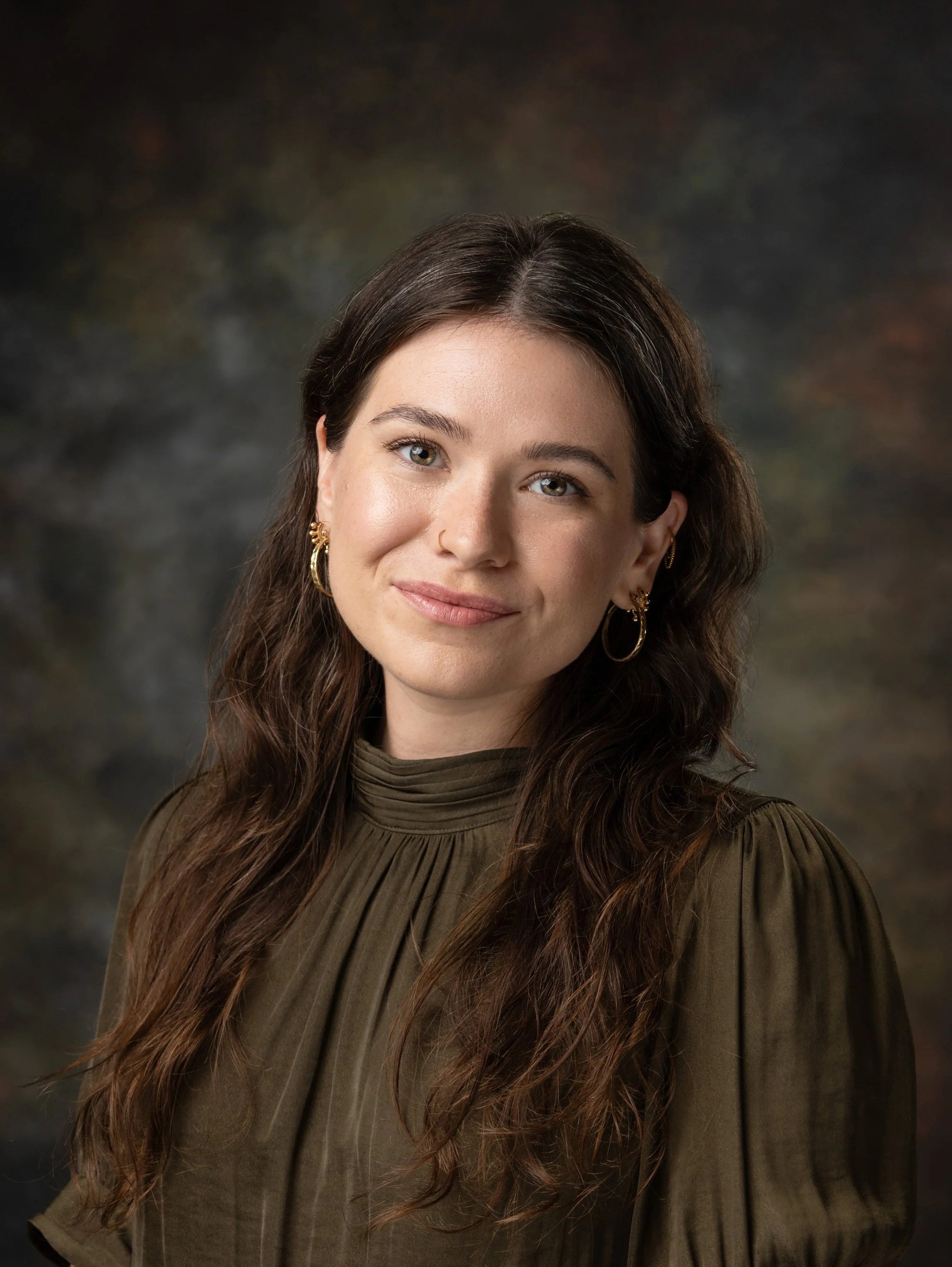 A woman with long wavy brown hair, light skin, smiling, wearing a dark button-up shirt, and a nose ring.