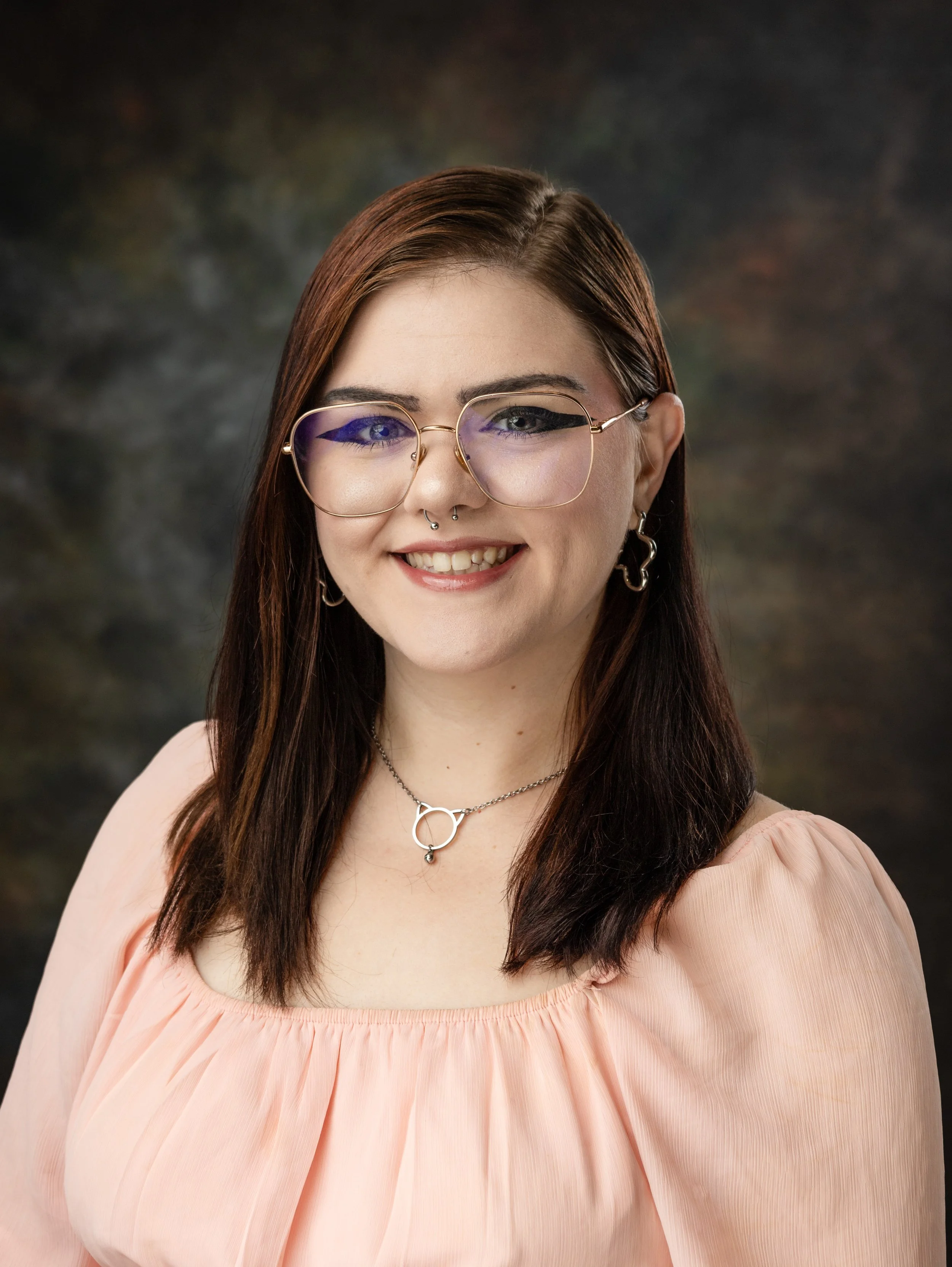 A woman with black and pink hair, wearing makeup, earrings, a choker, and a white cardigan, smiling at the camera.
