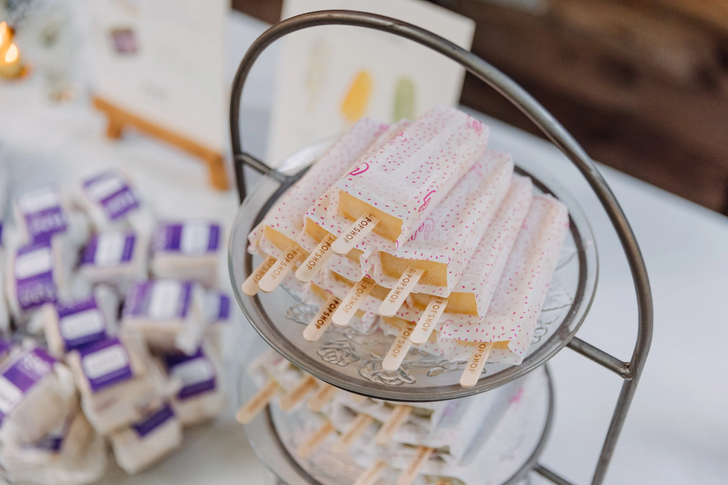 Assorted popsicles on a metal tiered display, including orange, yellow, and white varieties decorated with pink sprinkles, with other snack items in blurred background.