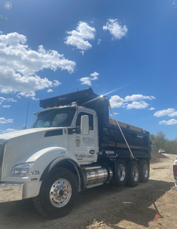A large white and black dump truck parked on dirt with a blue sky and scattered clouds above.