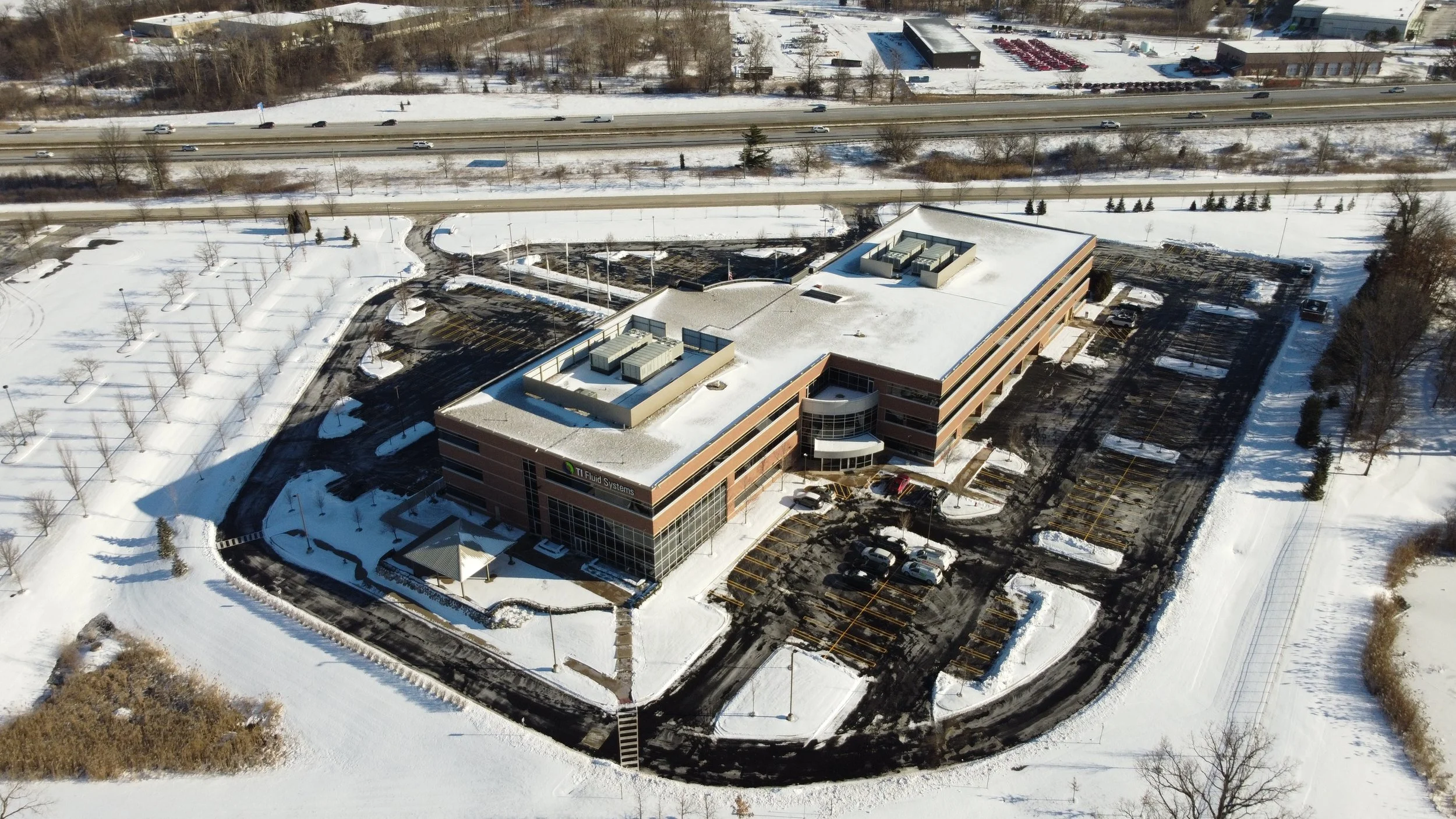 Aerial view of a large office building with a snow-covered parking lot and surrounding snow-covered landscape, including trees and roads.