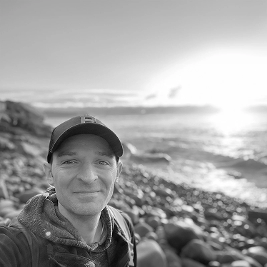 Black and white photo of Luke Pieczynski wearing a baseball hat  and hoody standing on a rocky beach at sunrise in Acadia National Park