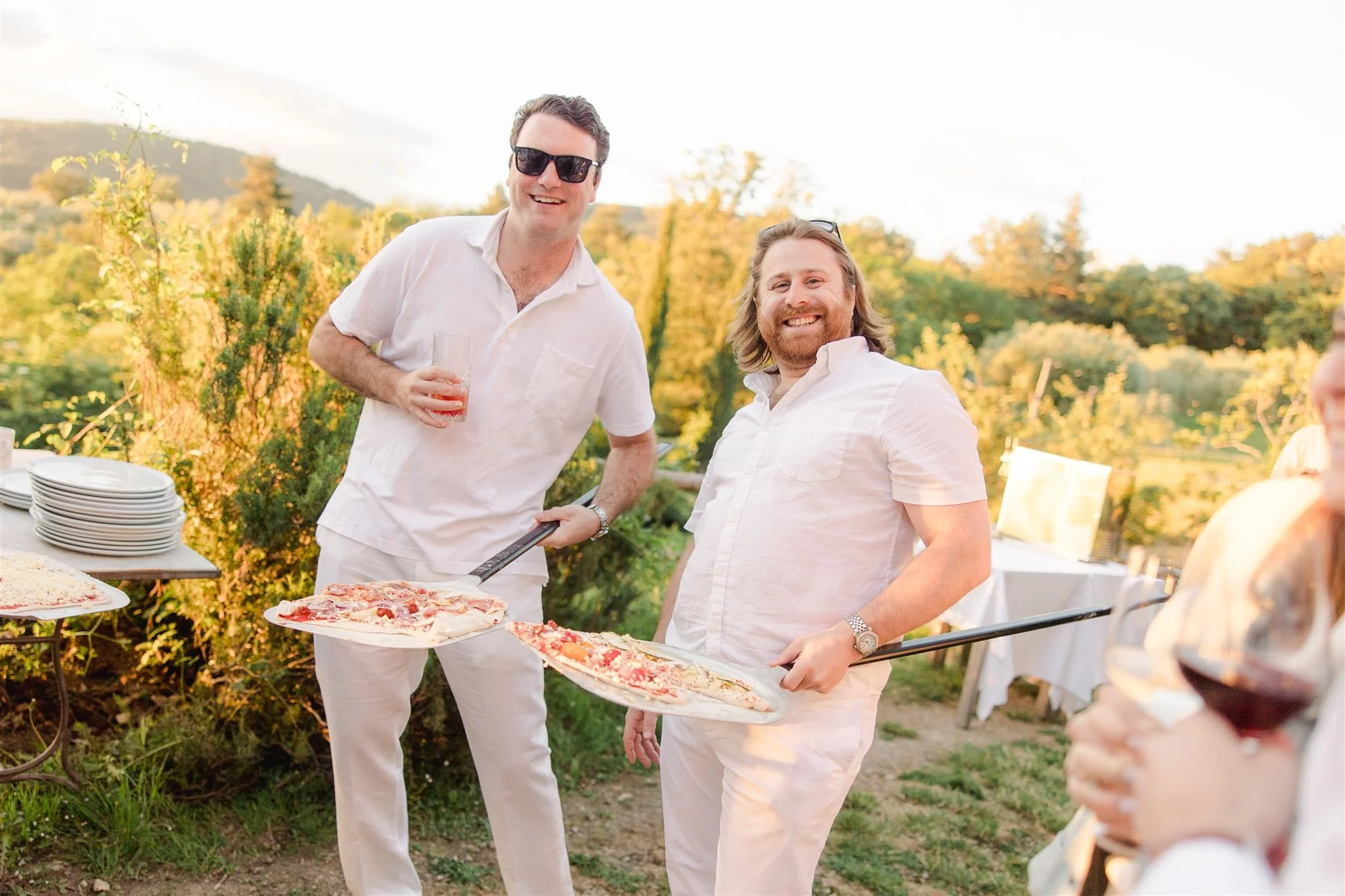 Two men in white shirts, smiling and holding pizza trays, socializing outdoors at a gathering during sunset at Castello di Ristonchi, a wedding venue in Tuscany for the welcome pizza party.