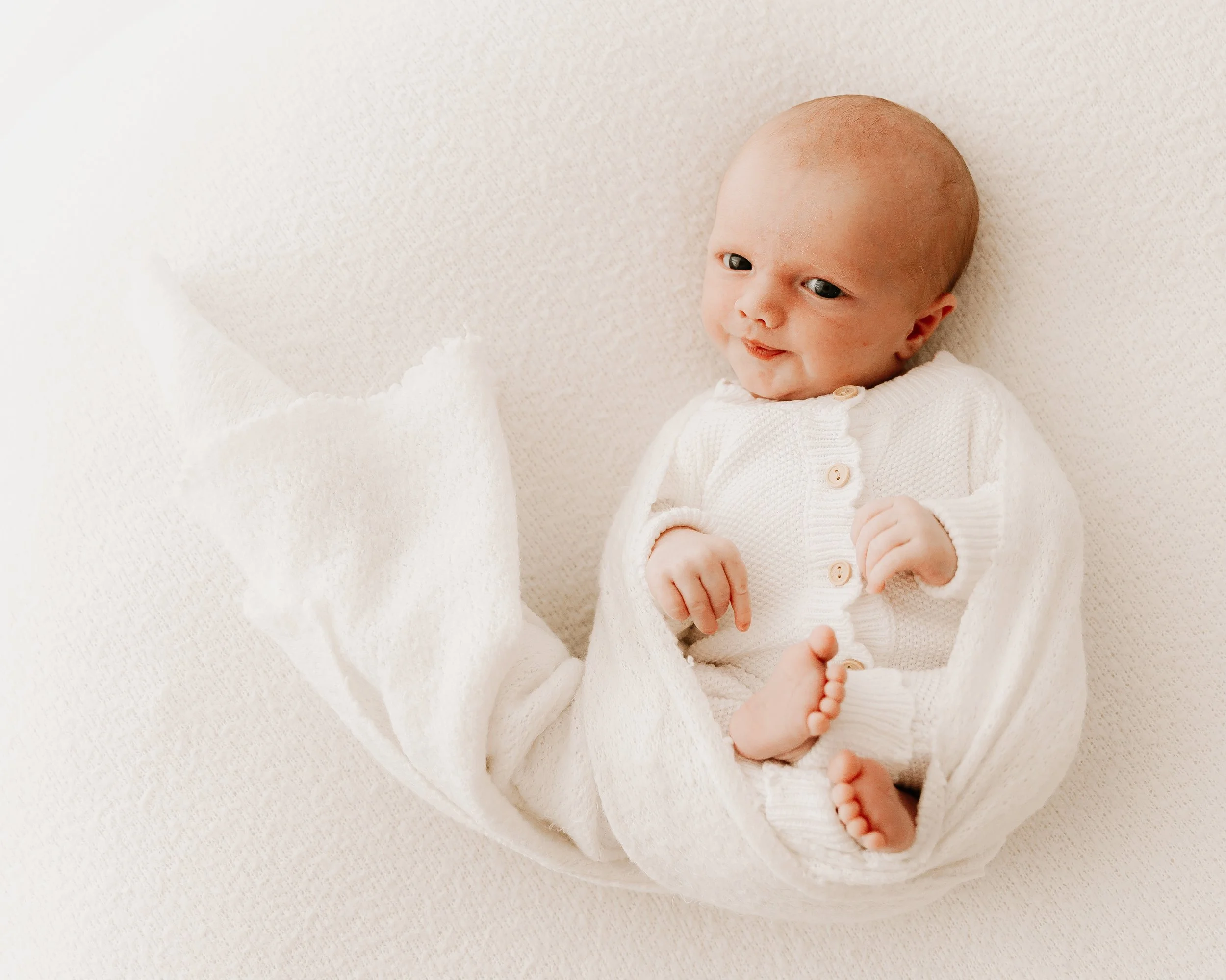 A smiling baby with a bald head, wearing a white knitted cardigan, lying on a soft white blanket, with a white blanket partially covering legs and feet.