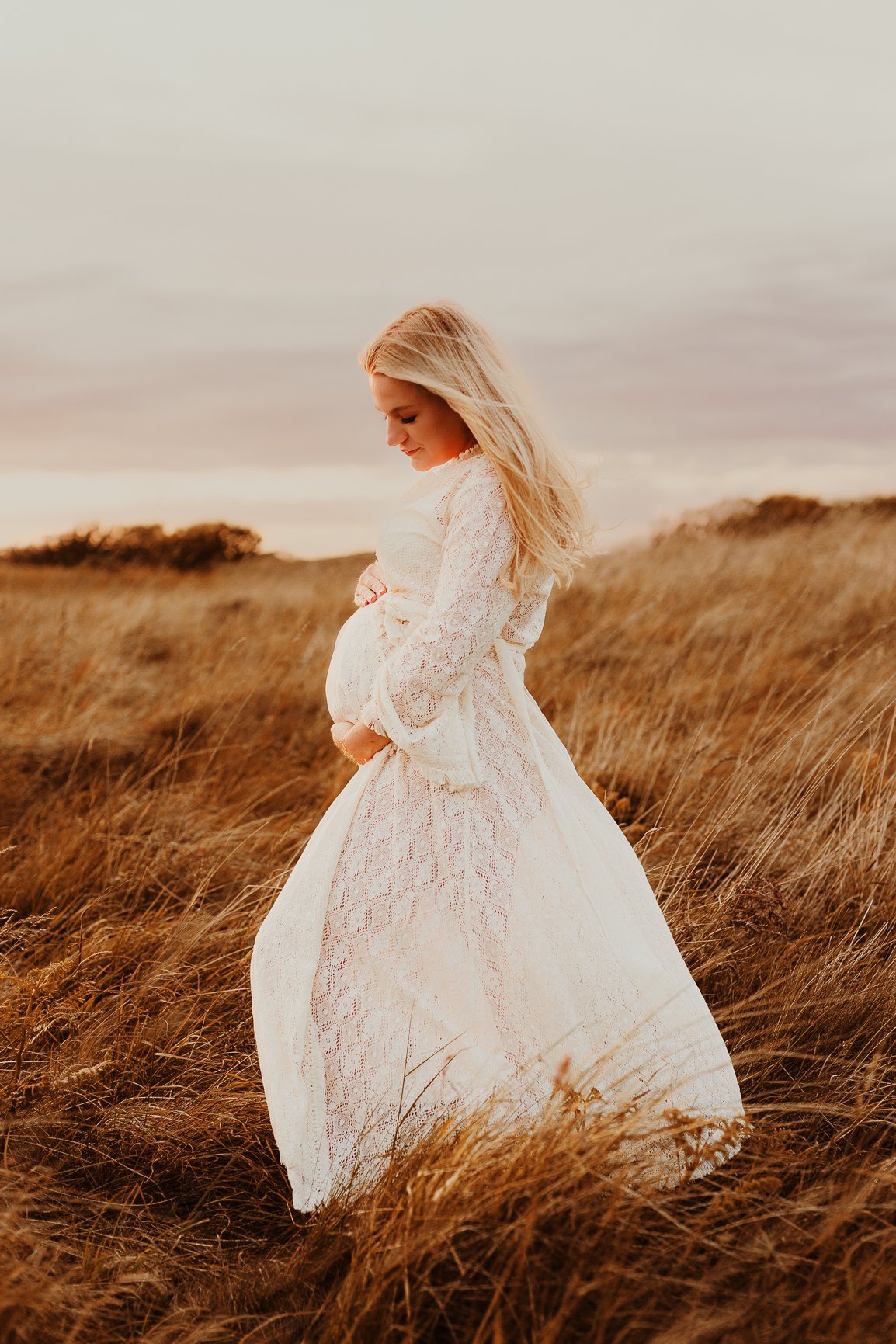 A pregnant woman in a long, white lace dress standing in a grassy field during sunset, gently holding her belly and looking down.
