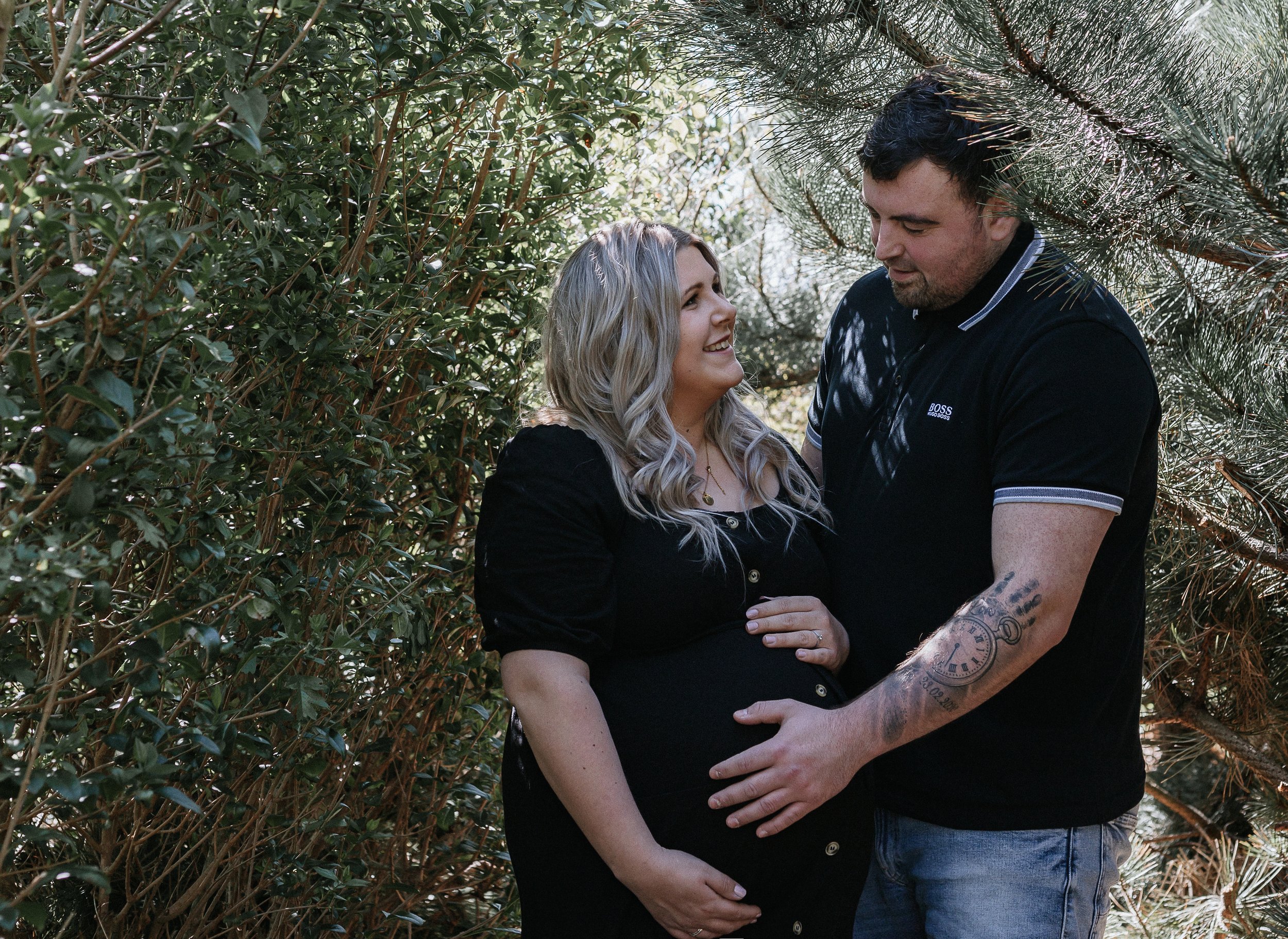 A pregnant woman smiling at a man amidst trees and foliage.