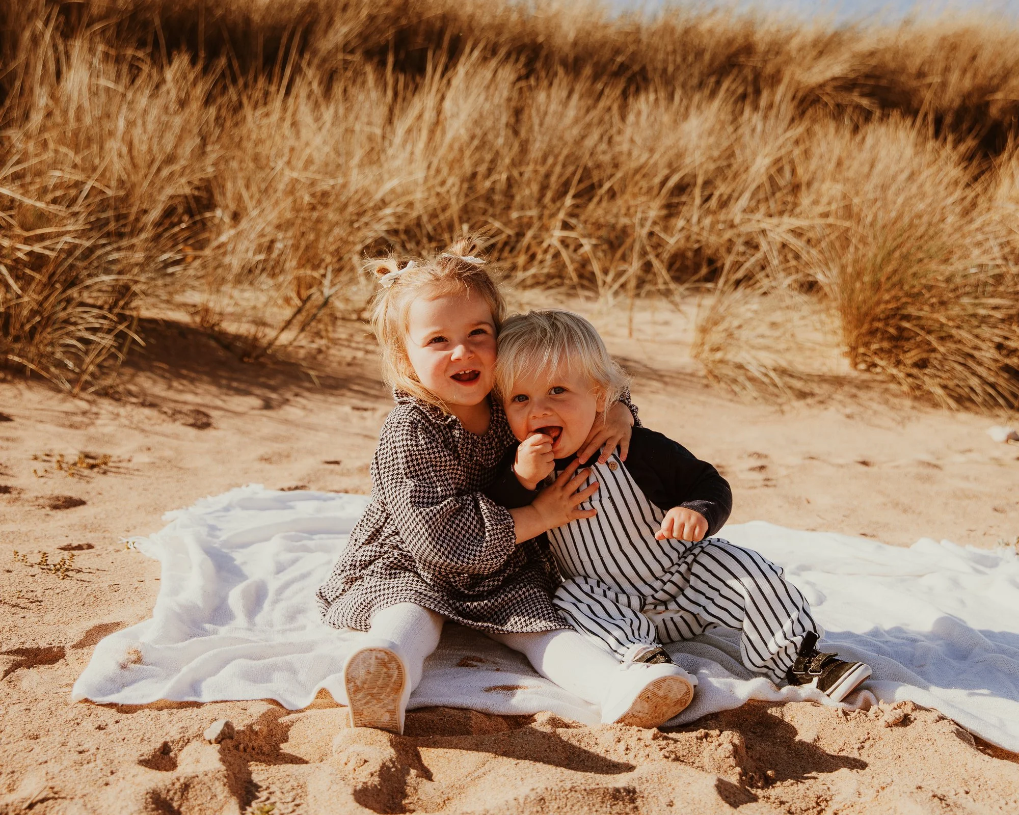 Two young children, a girl and a boy, sitting on a white blanket on a sandy beach, hugging and smiling surrounded by tall sand dunes and beach grass.