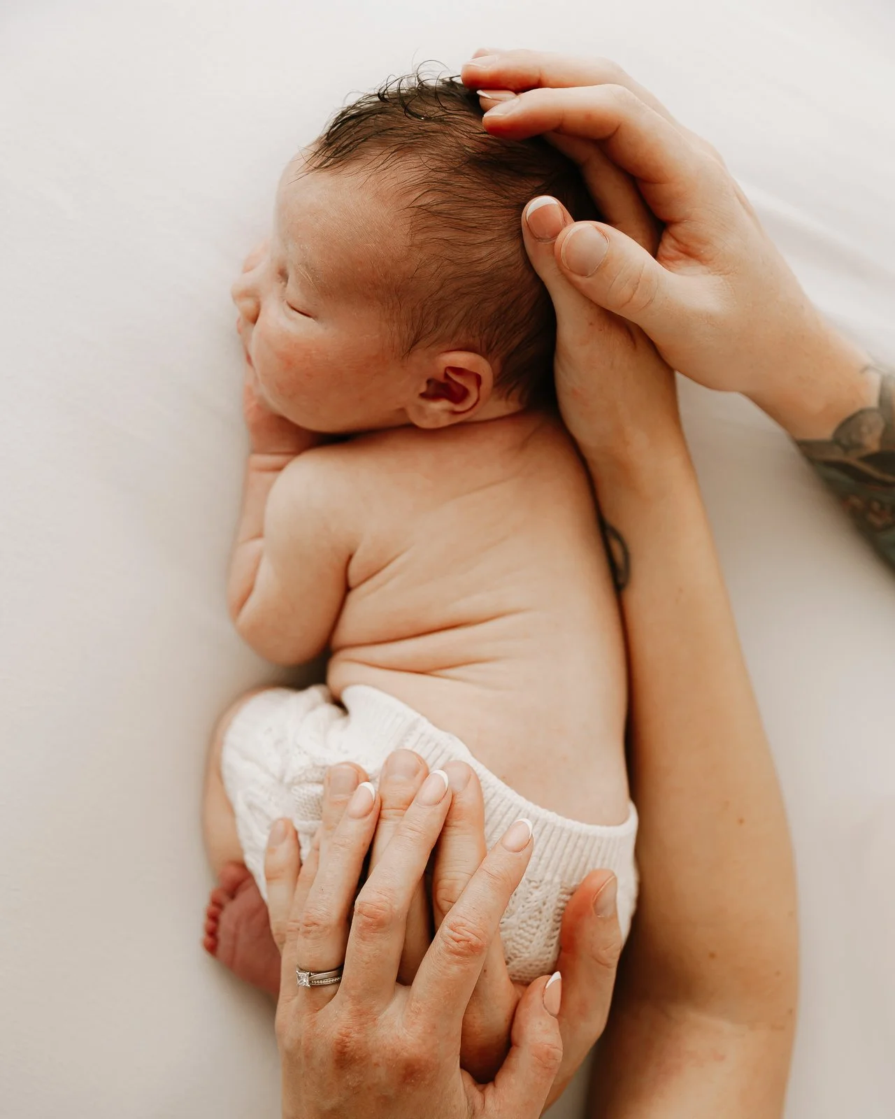 A newborn baby sleeping on a white surface, being cradled gently by adult hands, with a visible ring on the adult's finger.