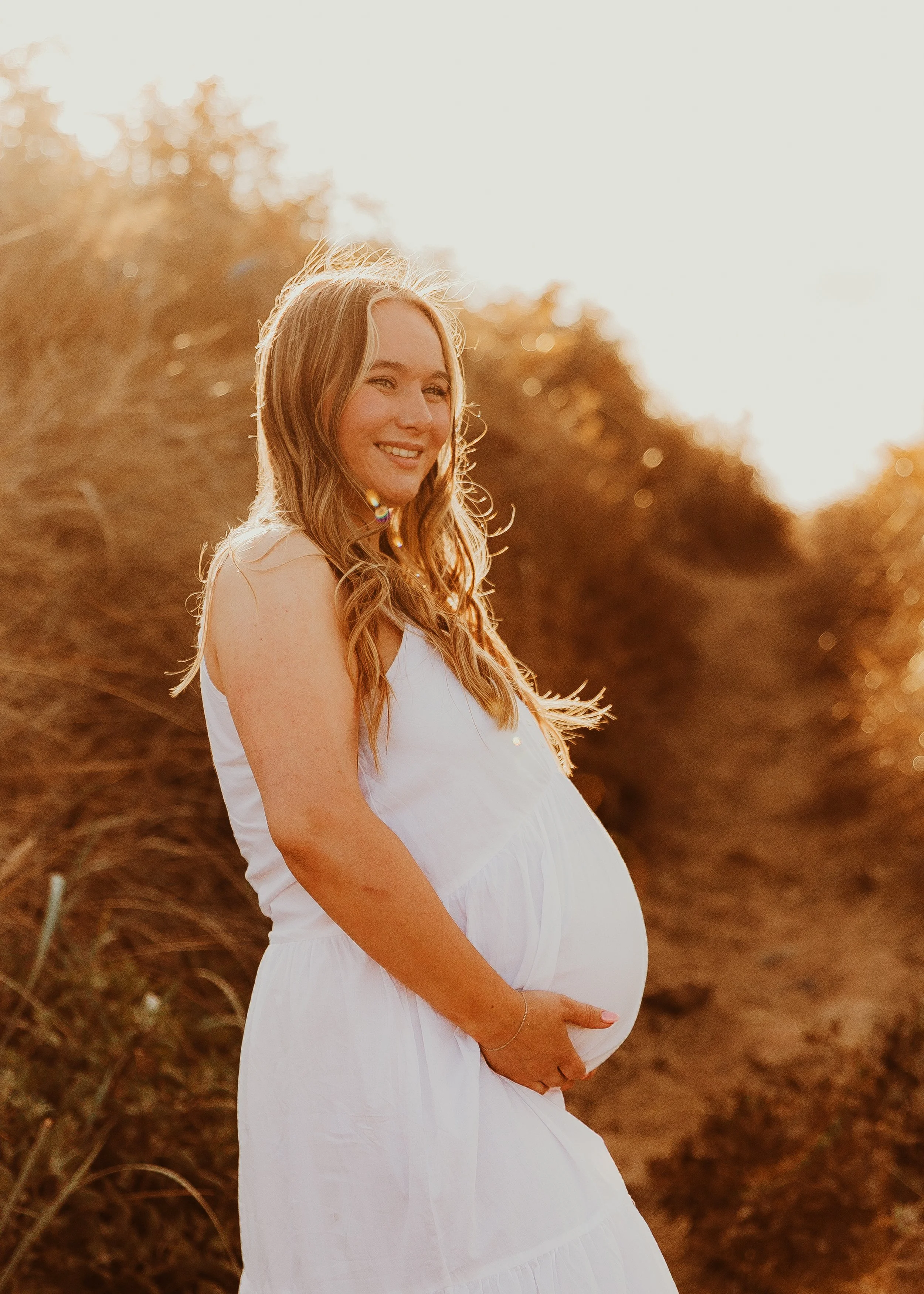 A pregnant woman with long wavy hair in a white sleeveless dress stands outdoors in a field of tall plants during sunset, smiling and gently holding her baby bump.