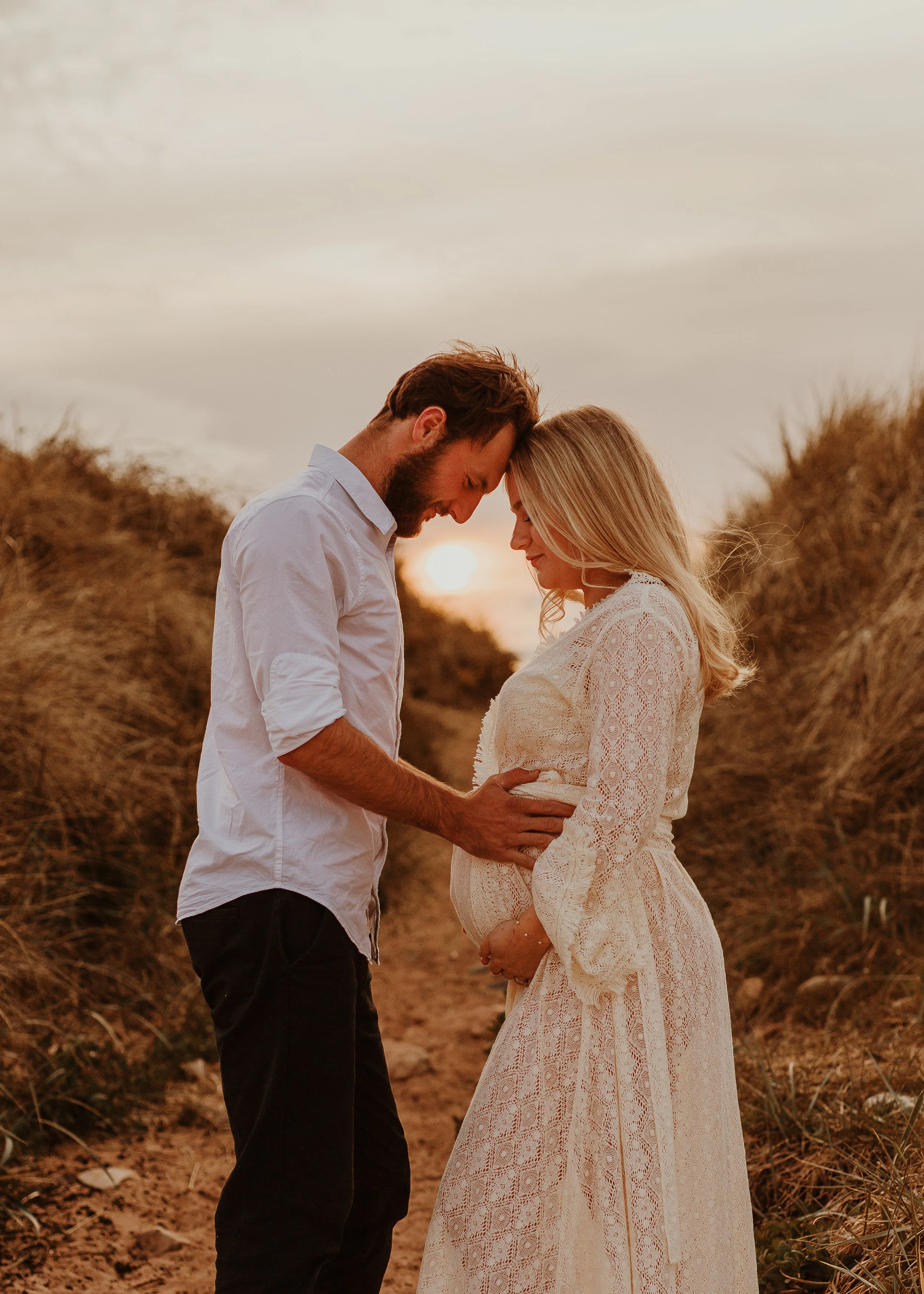 A couple stands close together on a sandy path with tall grass, with the sun setting behind them. The woman is pregnant, and they are gently touching her belly with their foreheads touching, sharing a tender moment.
