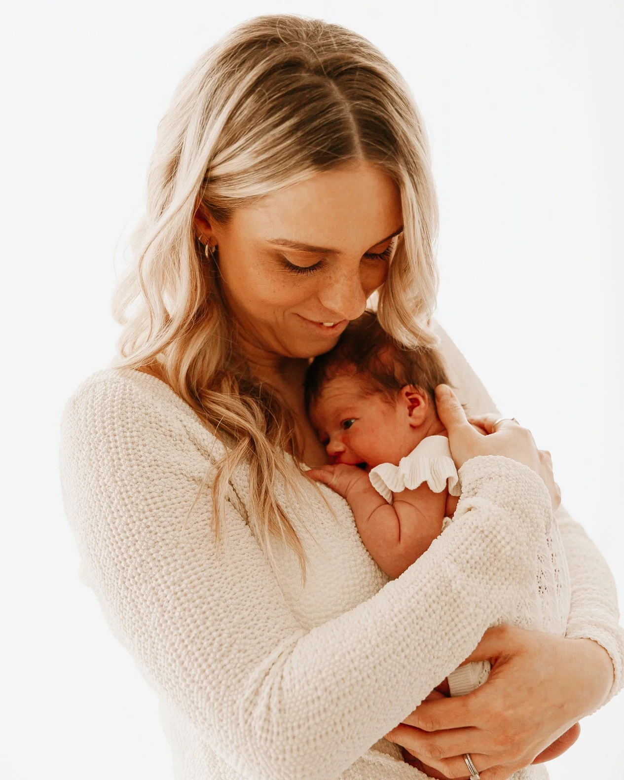 A woman holding a newborn baby close to her chest, both smiling and looking down, with a plain white background.