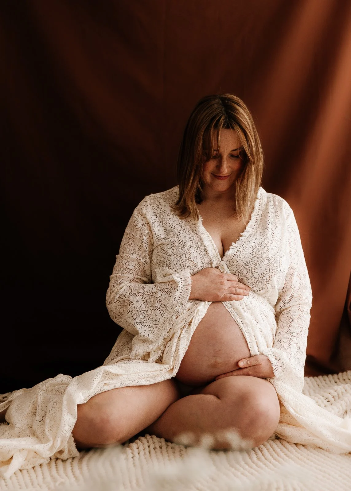 A pregnant woman sitting on a bed with a cream knit blanket, wearing a white lace robe, looking down at her belly, smiling gently.
