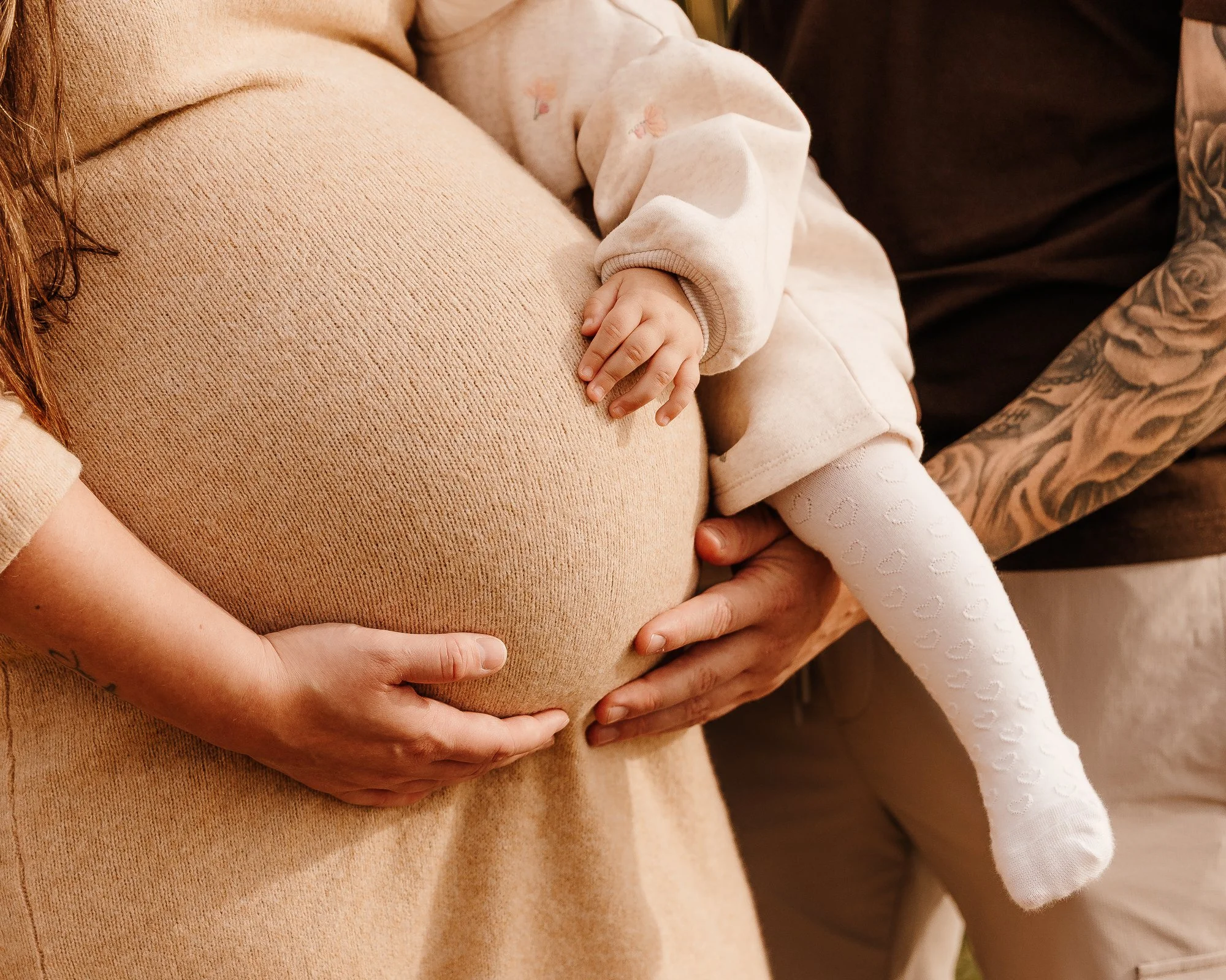 A pregnant woman and a young girl, likely her daughter, touch her pregnant belly while an adult man's hands support her waist in the background.