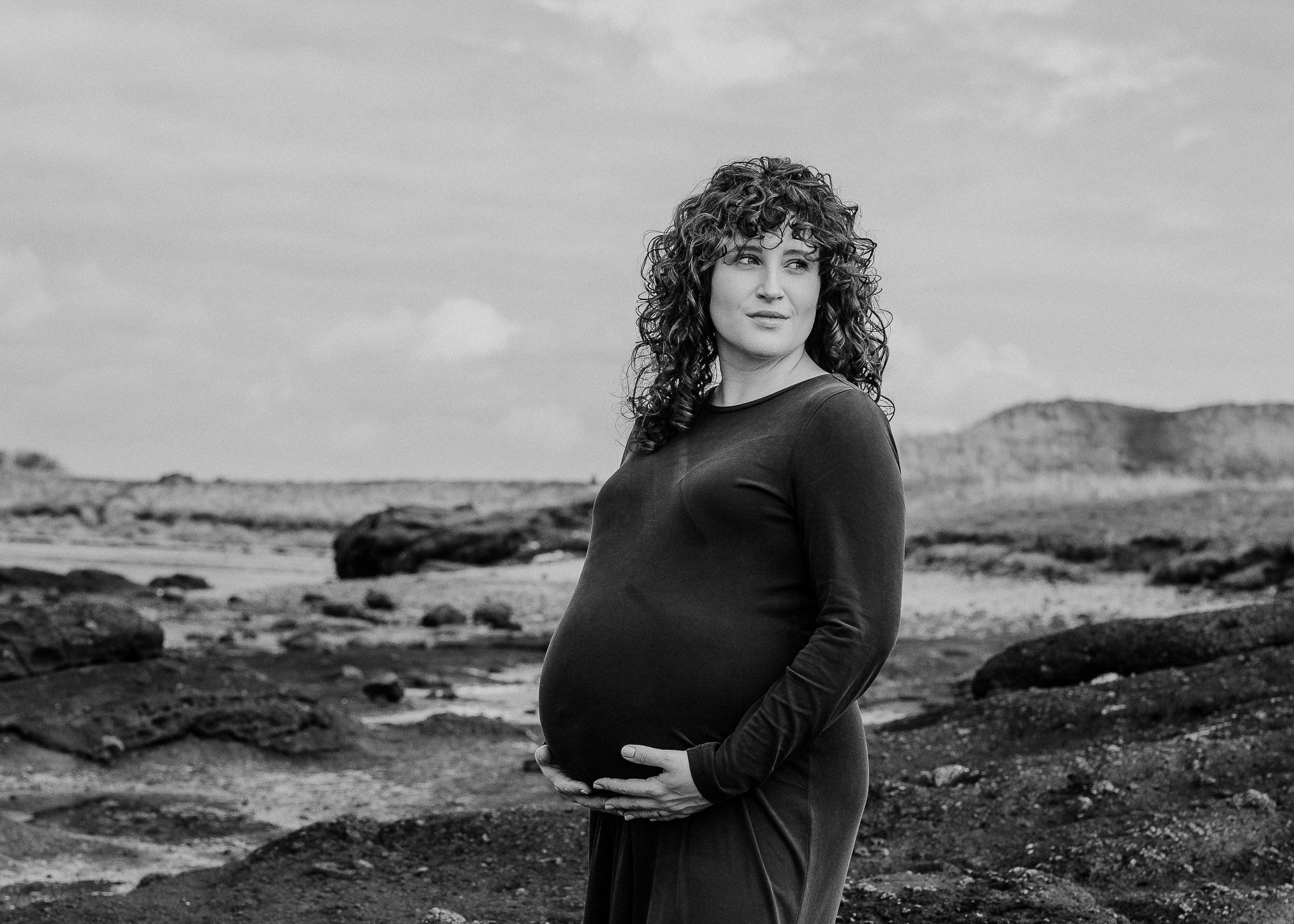 A pregnant woman standing outdoors on rocky terrain, holding her belly and looking to the side, with a landscape and hills in the background. The photo is in black and white.
