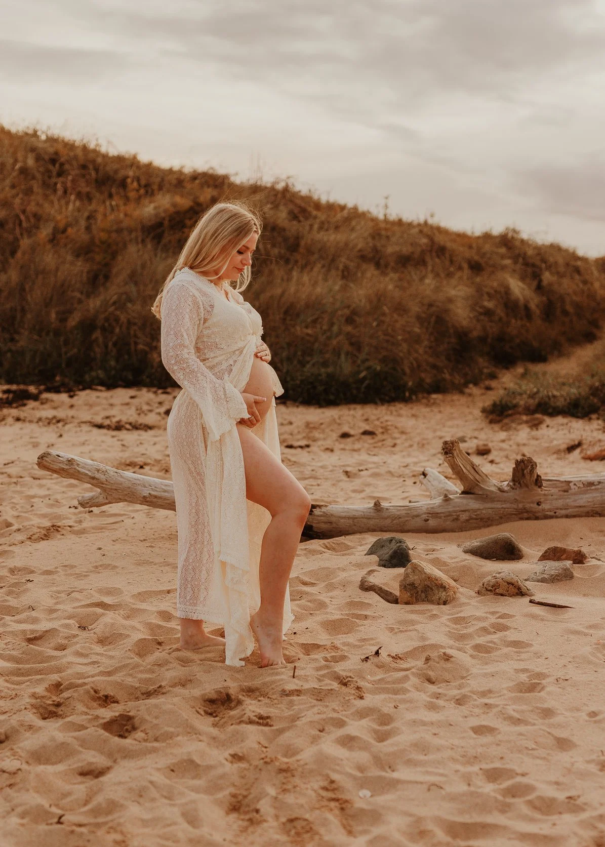 Pregnant woman in a white lace dress standing on a sandy beach with driftwood and rocks, looking at her belly, with grassy dunes and a cloudy sky in the background.