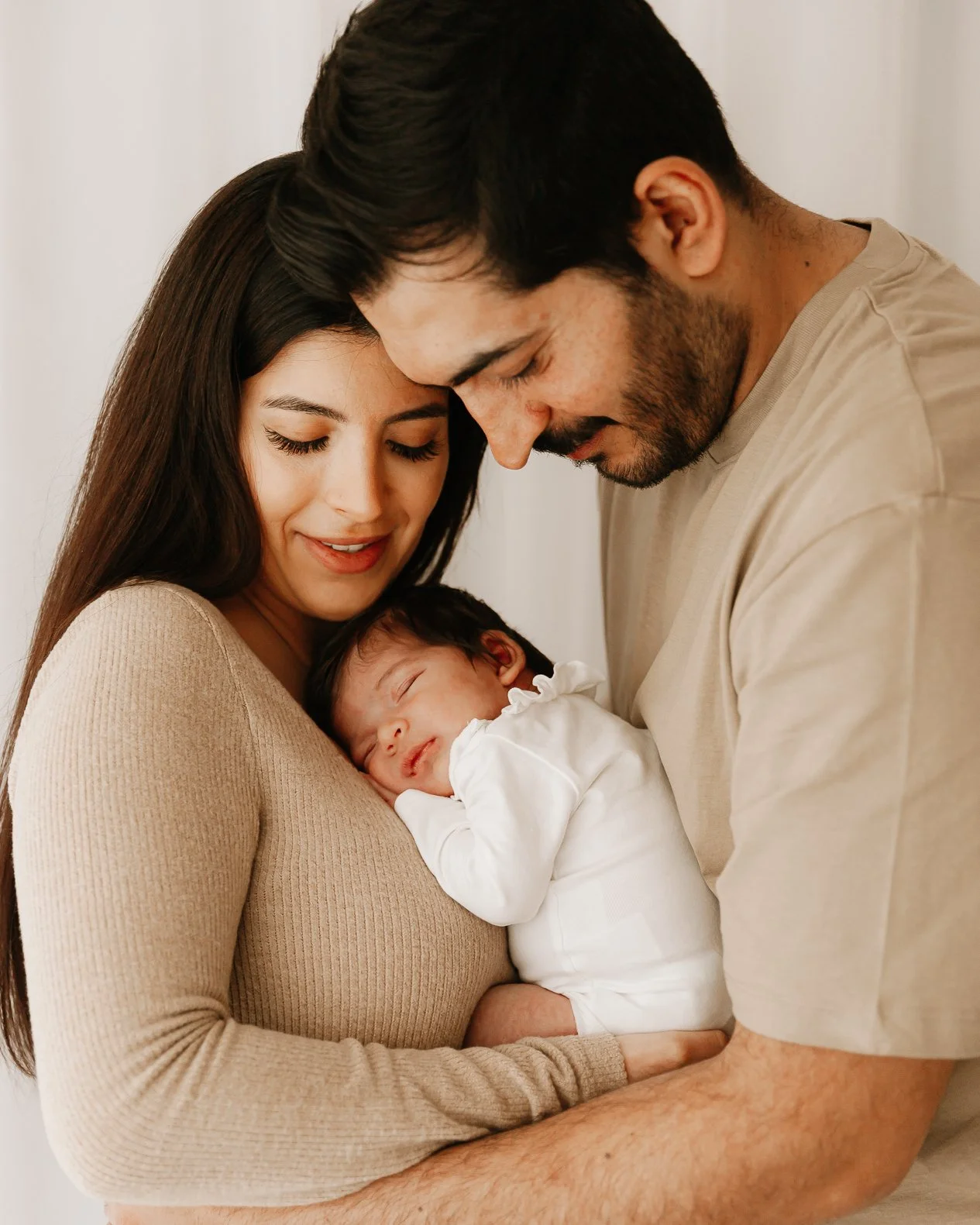 A family of three, a man and a woman holding their sleeping baby, all with their foreheads touching, in a tender moment.