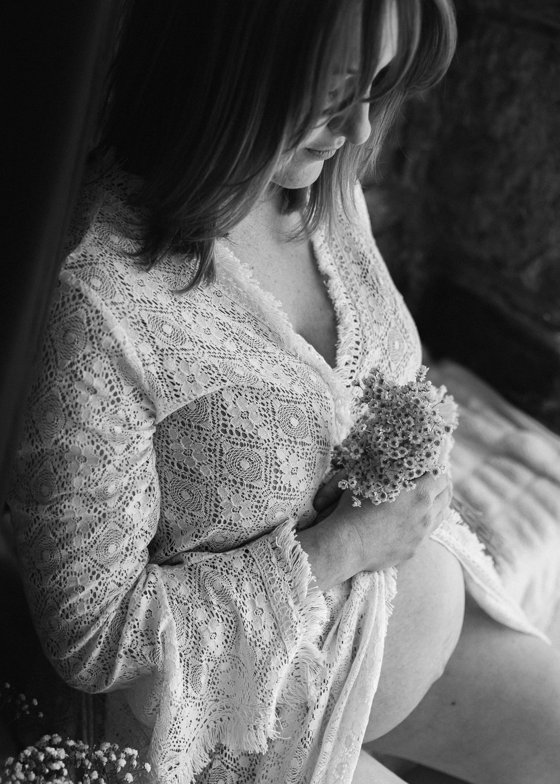 A pregnant woman in lace clothing holding a small bouquet of flowers, looking down at her belly.